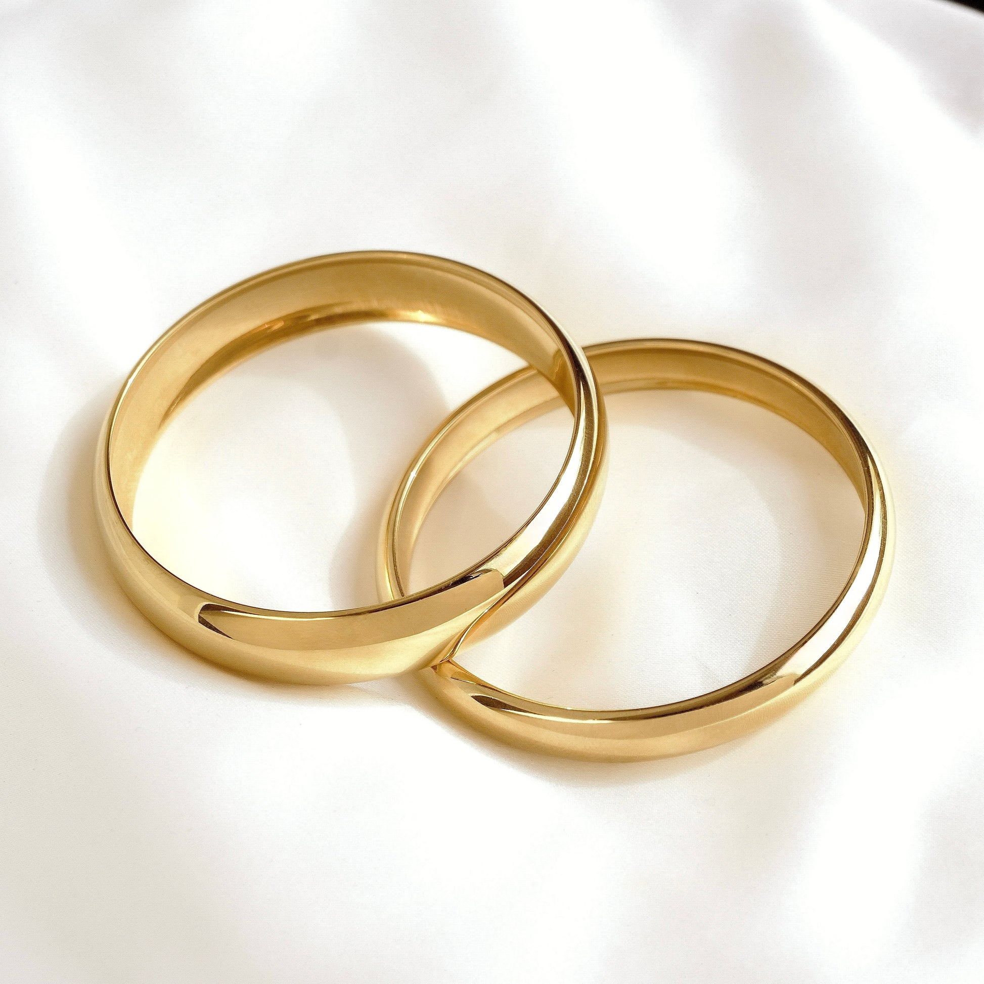 Two polished gold bangle bracelets resting on a soft white fabric background.