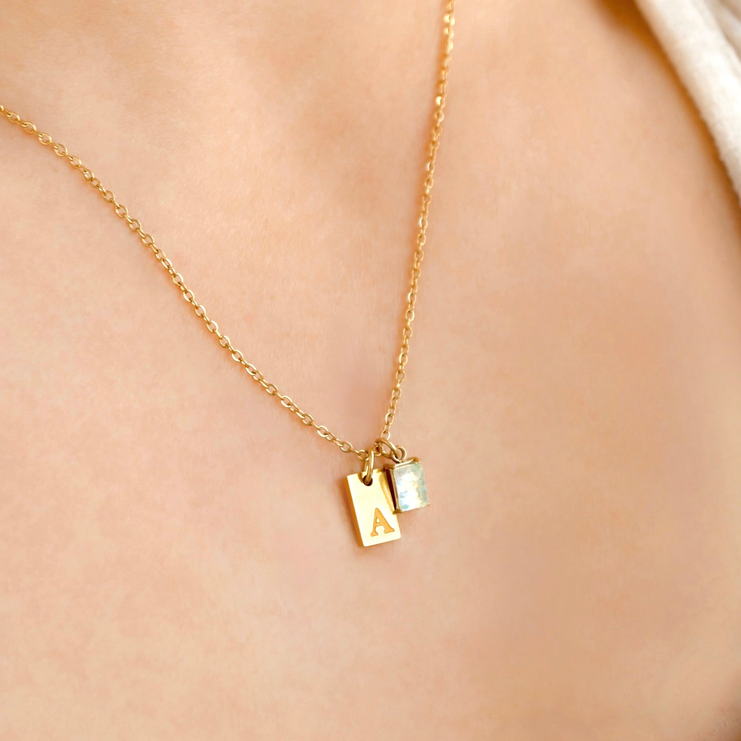 Gold necklace with a small tag initial and opal birthstone on a person's neck against a beige background