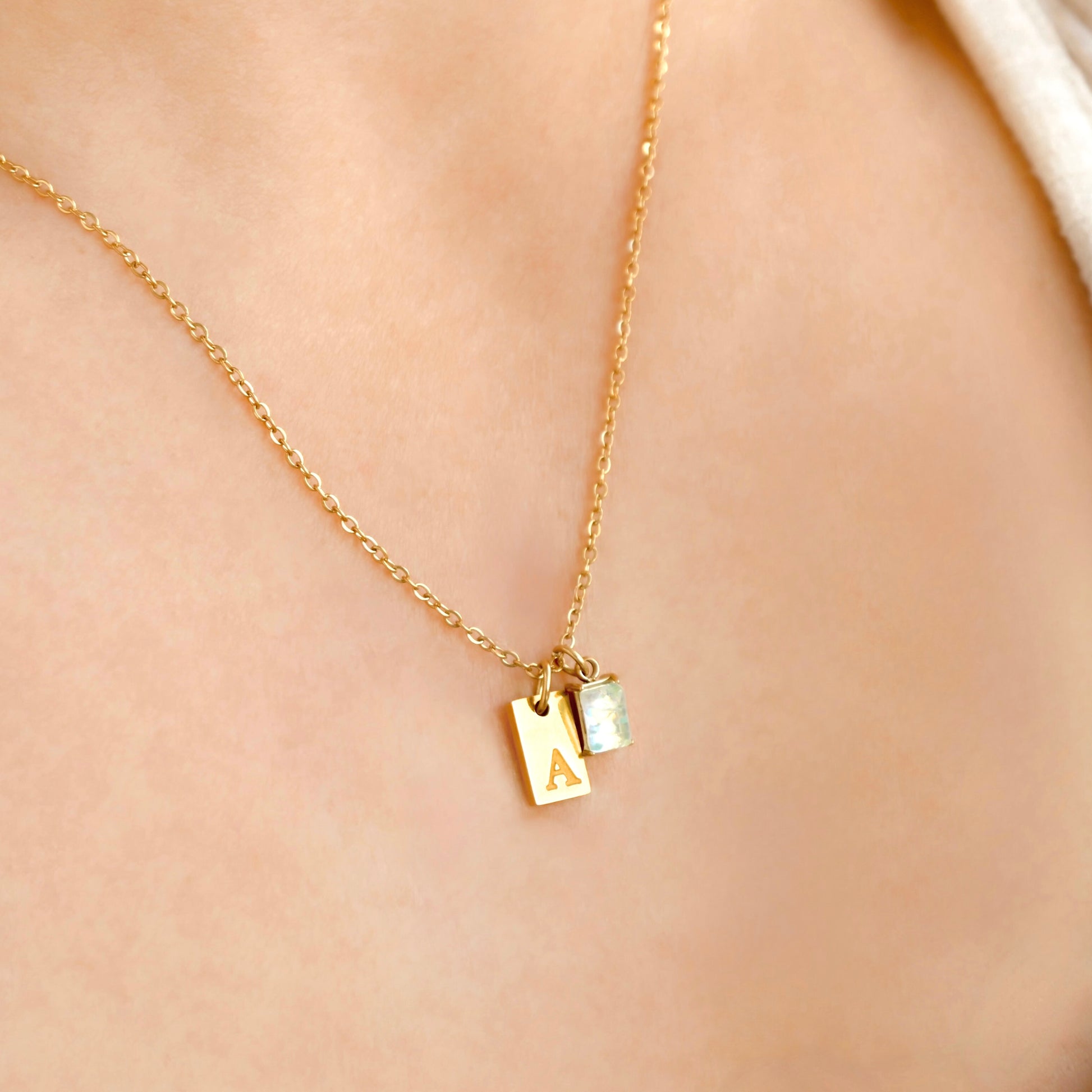 Gold necklace with a small tag initial and opal birthstone on a person's neck against a beige background