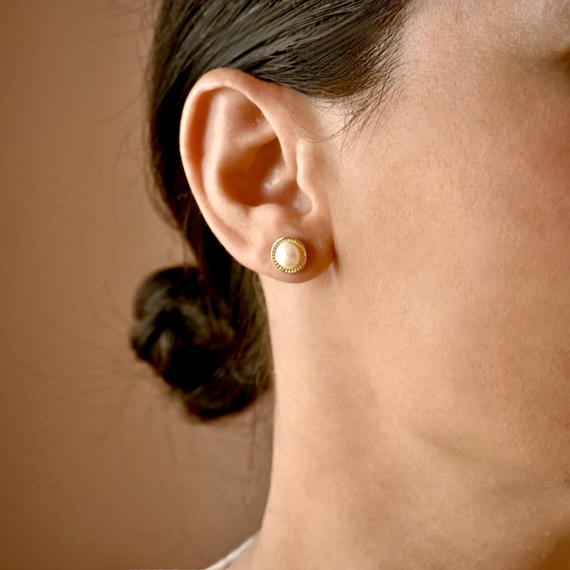 A close-up side profile of a woman's ear wearing a small round gold stud earring with a cream-colored pearl-like center, against a warm brown background.