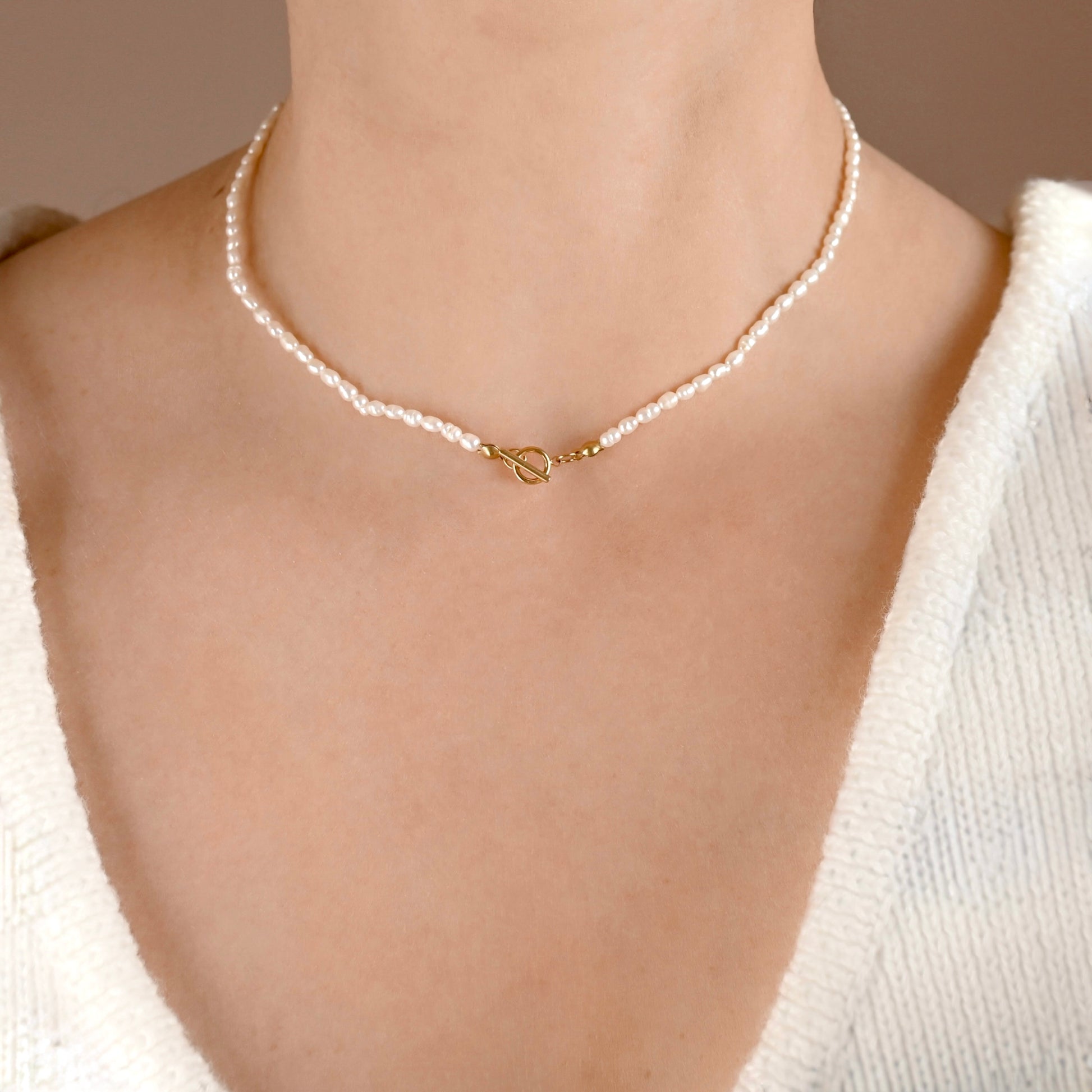 A close-up of a person's neck and upper chest wearing a delicate pearl choker necklace with a small gold clasp, paired with a white knit sweater.
