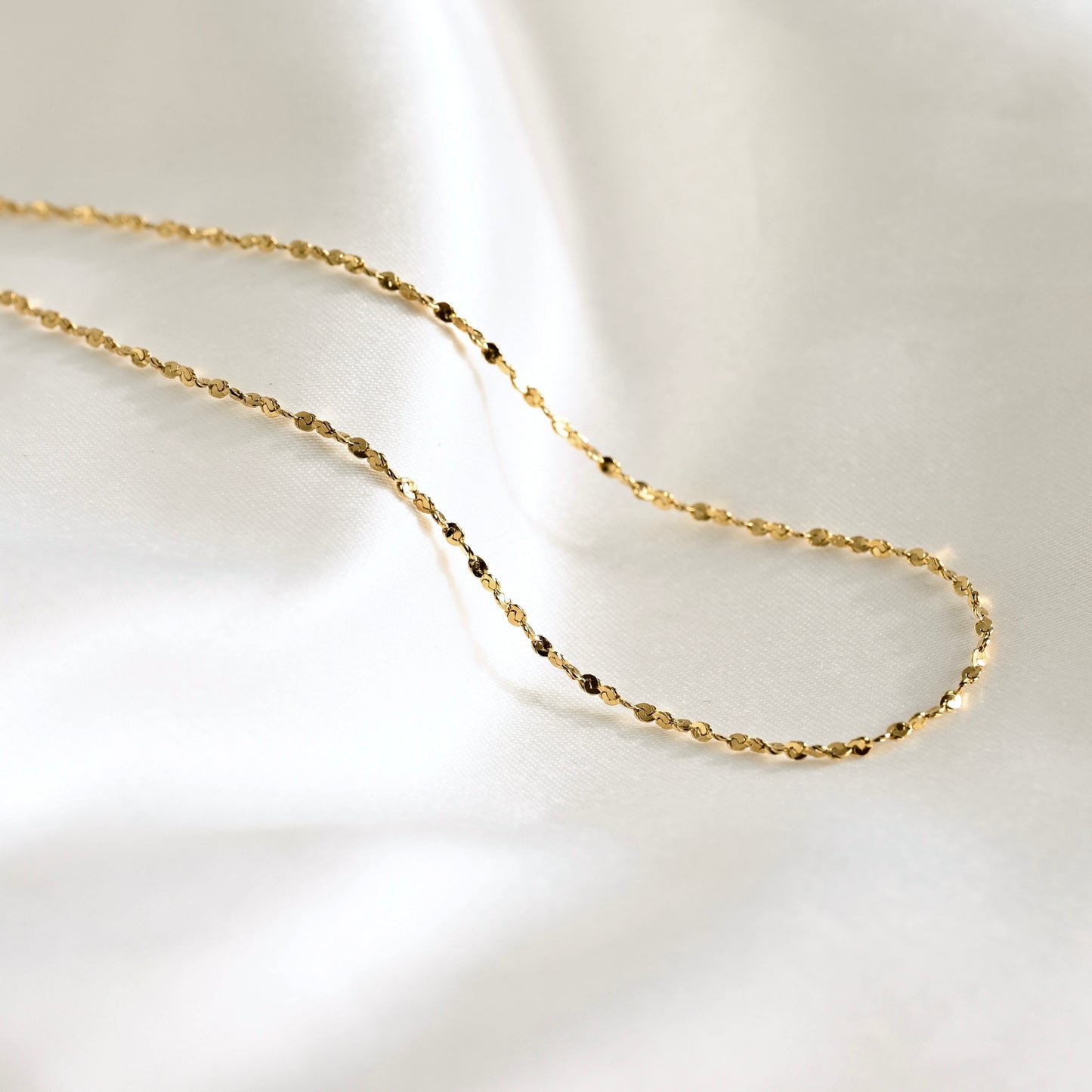 A delicate gold chain necklace with small, faceted beads, draped elegantly on a soft, white satin fabric background.