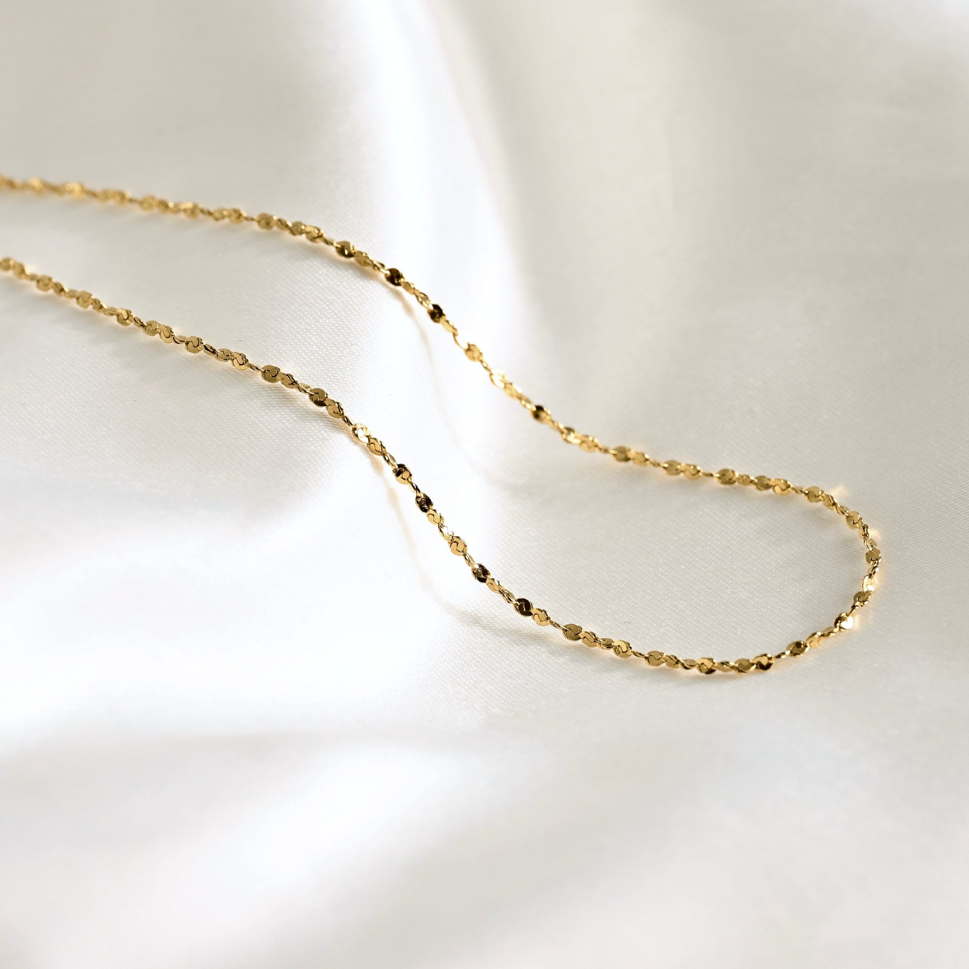 A delicate gold chain necklace with small, faceted beads, draped elegantly on a soft, white satin fabric background.