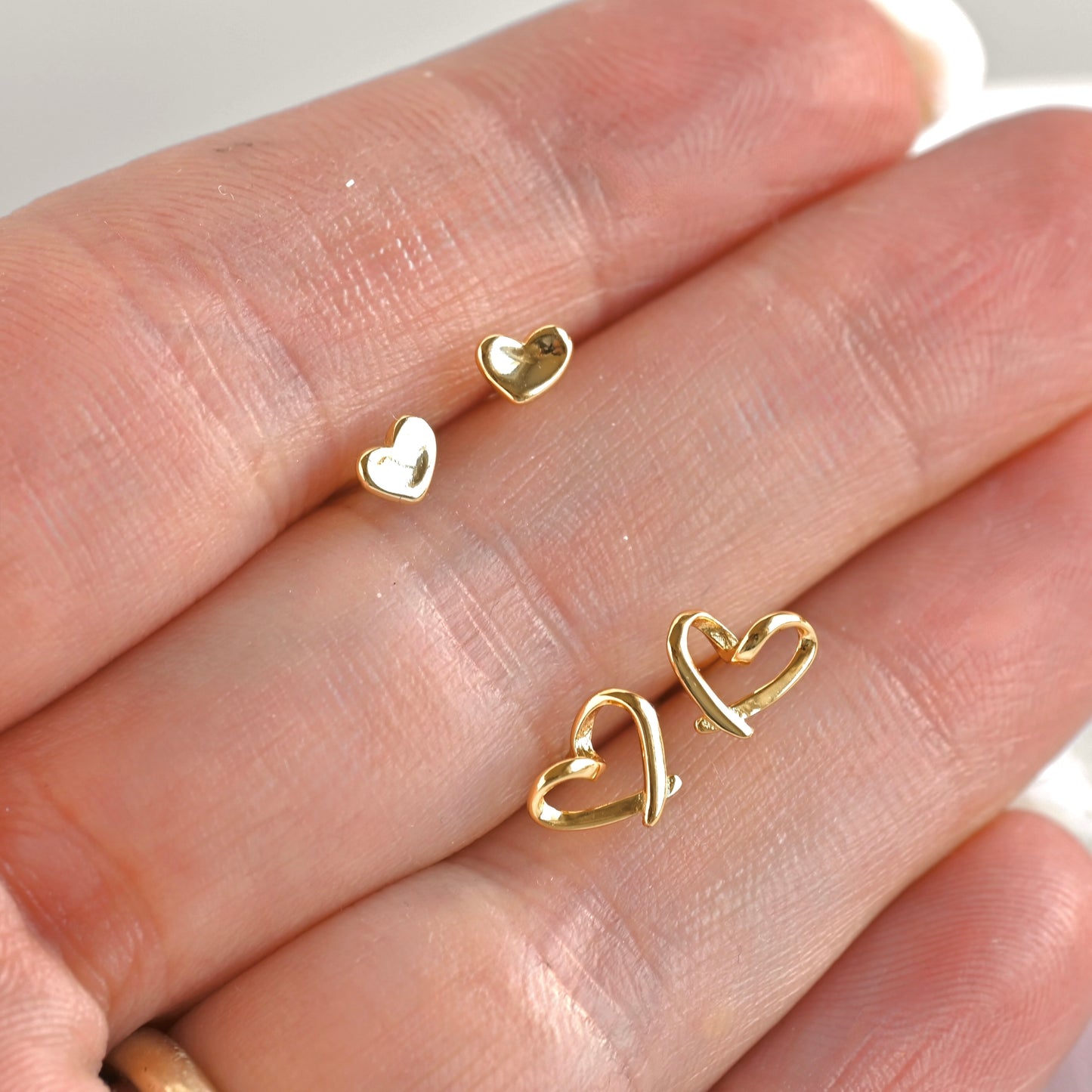 Three small gold heart-shaped stud earrings displayed on a person's fingers against a soft light background.