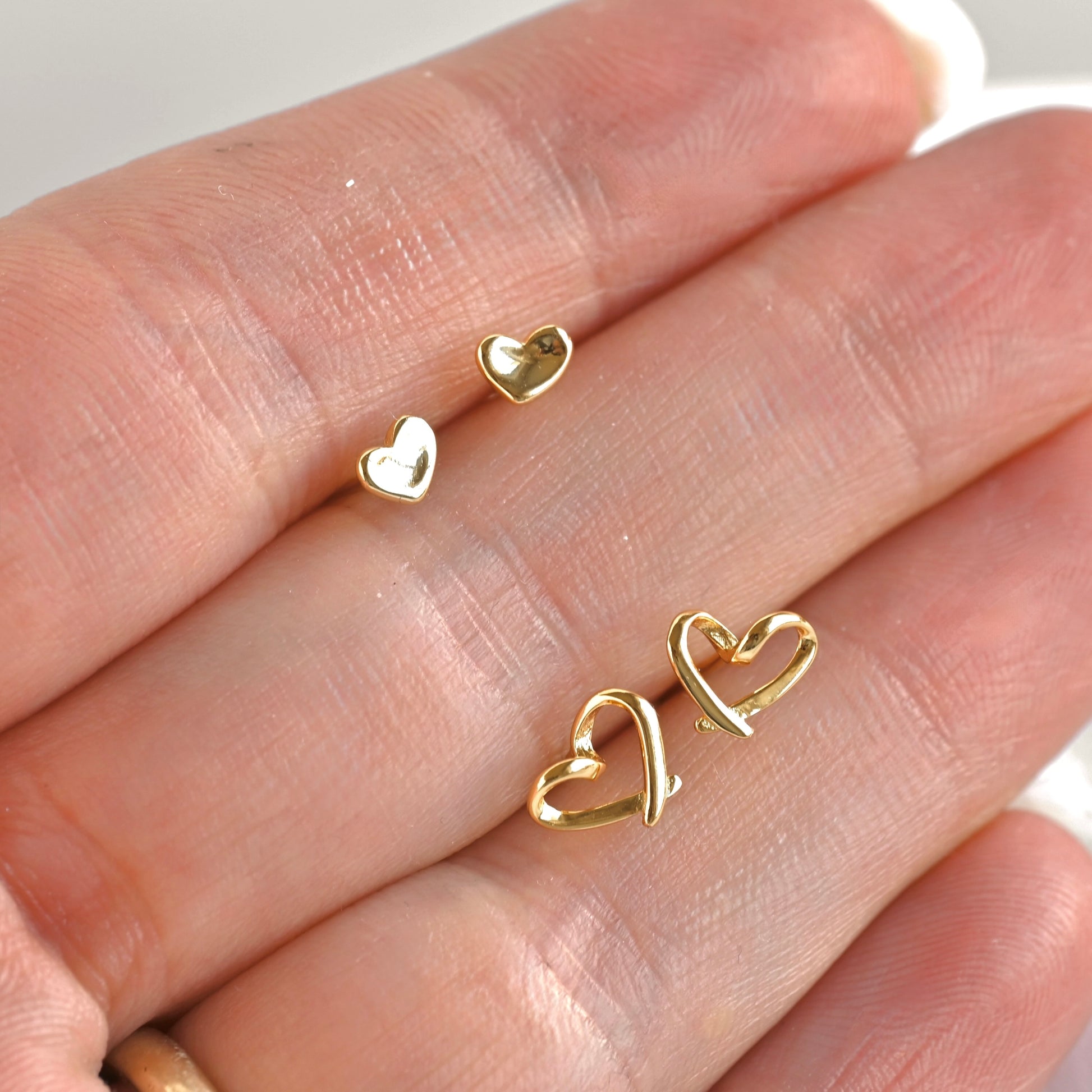 Three small gold heart-shaped stud earrings displayed on a person's fingers against a soft light background.