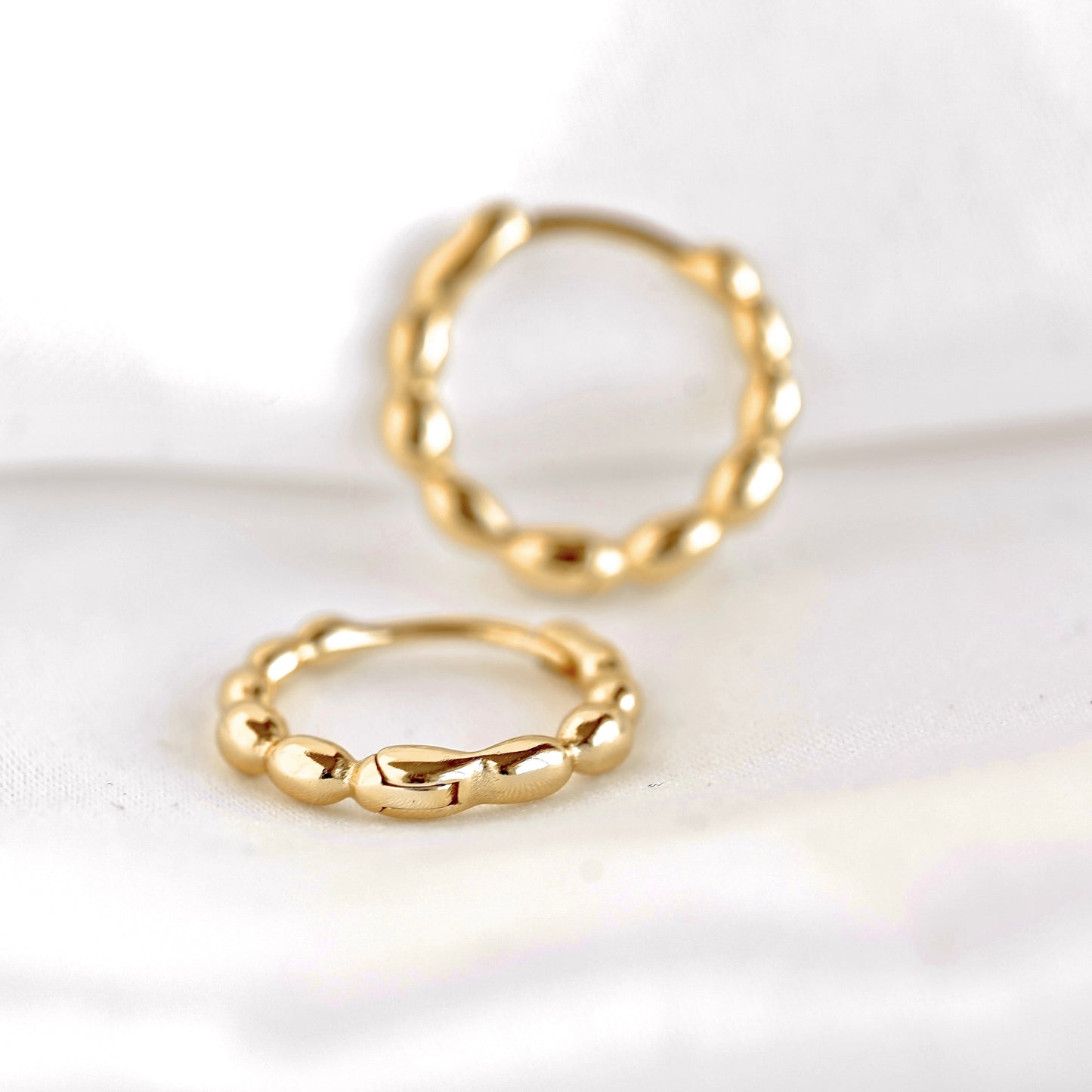 Two gold-toned twisted beaded earrings on a soft white fabric background.