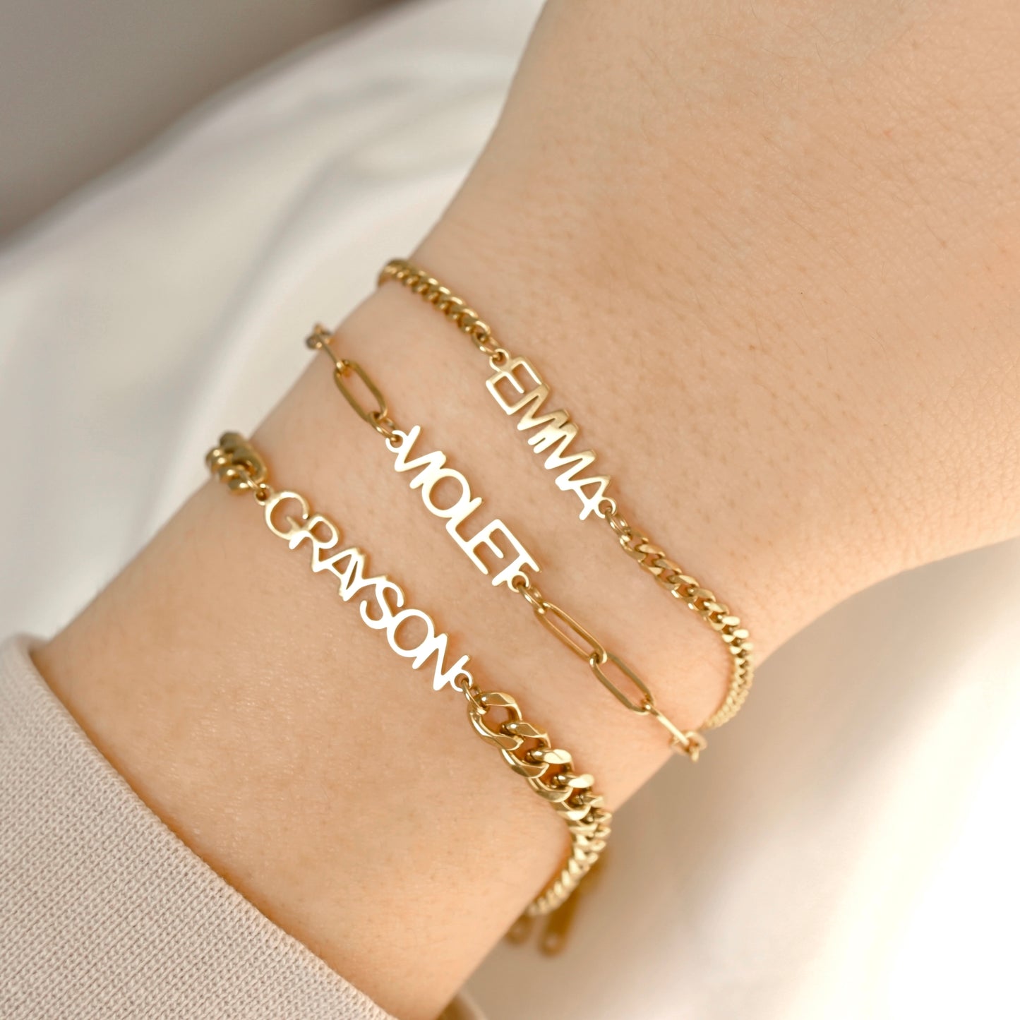 Three personalized gold name bracelets worn on a person's wrist, featuring link chains and script lettering with the names Emma, Violet, and Grayson.