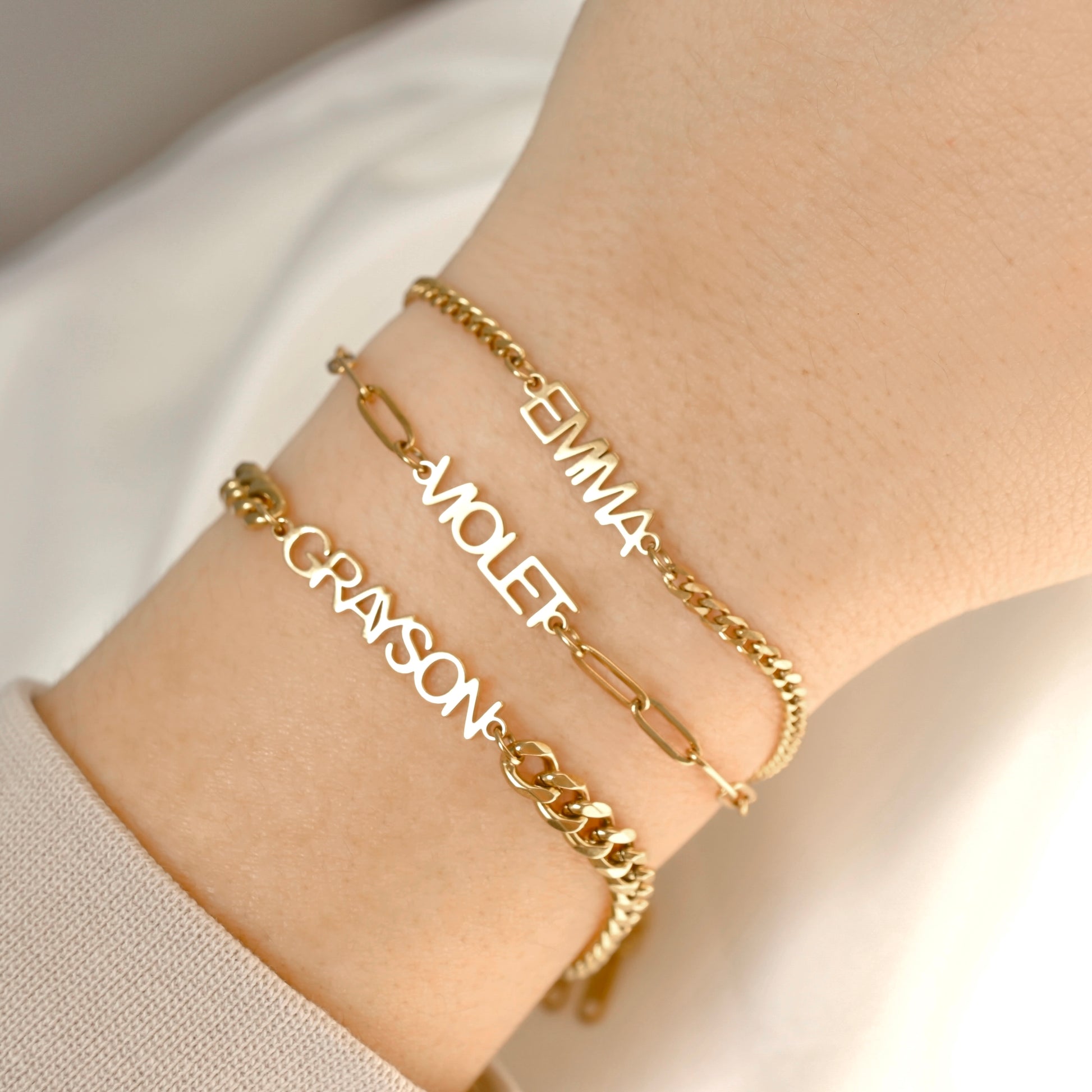 Three personalized gold name bracelets worn on a person's wrist, featuring link chains and script lettering with the names Emma, Violet, and Grayson.