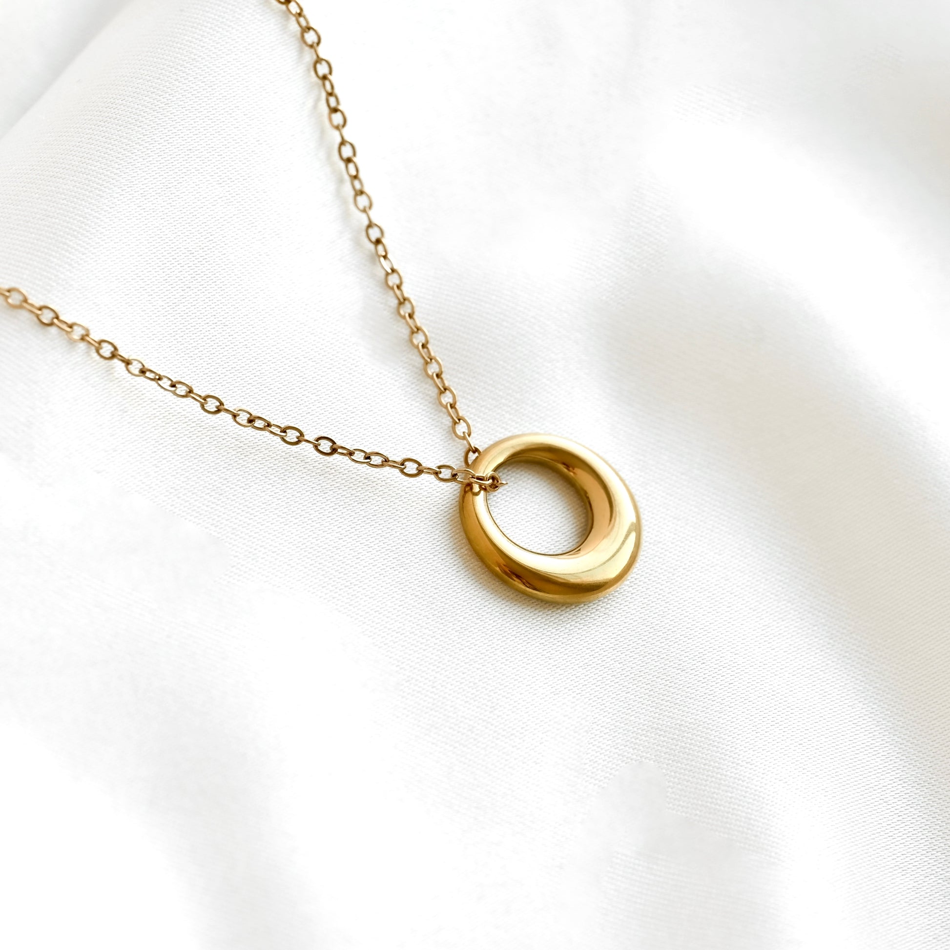 A gold chain necklace with a polished, open circular pendant, displayed on a soft, white textured fabric background.