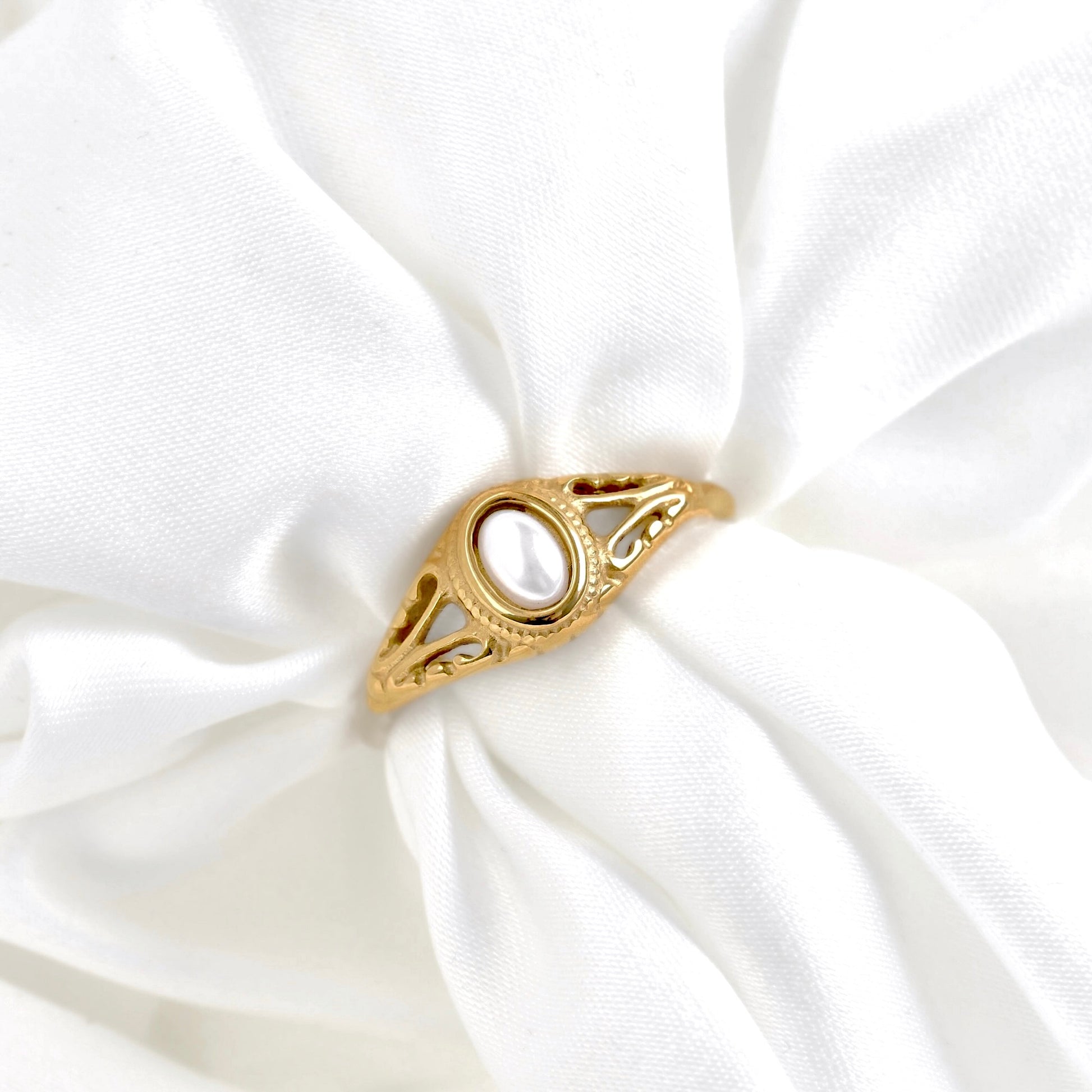 A gold ring with an oval white pearl-like center stone, displayed on a soft white satin fabric.
