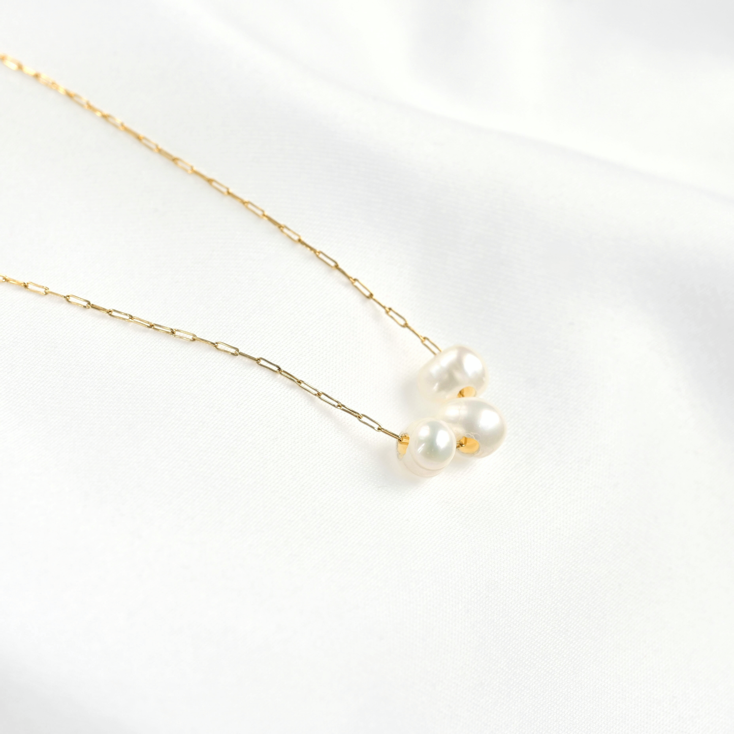 A delicate gold chain necklace featuring three small white freshwater pearls arranged in a cluster, displayed on a soft white fabric background.