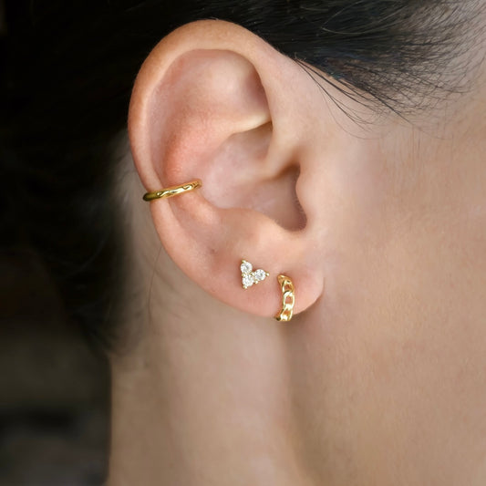 Close-up of an ear wearing gold curb link huggies, clover stud earrings and a thin hammered ear cuffs, all in sterling silver.