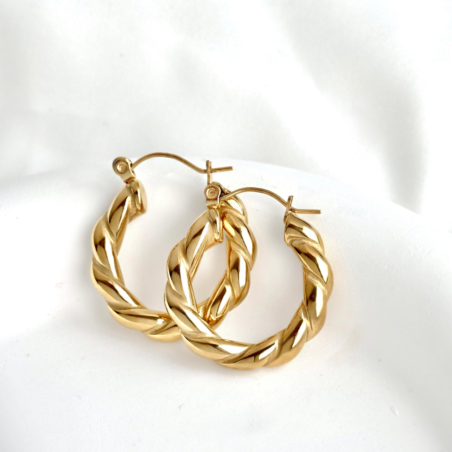 A pair of gold hoop earrings with a twisted, braided rope-like design, displayed on a soft white fabric background.