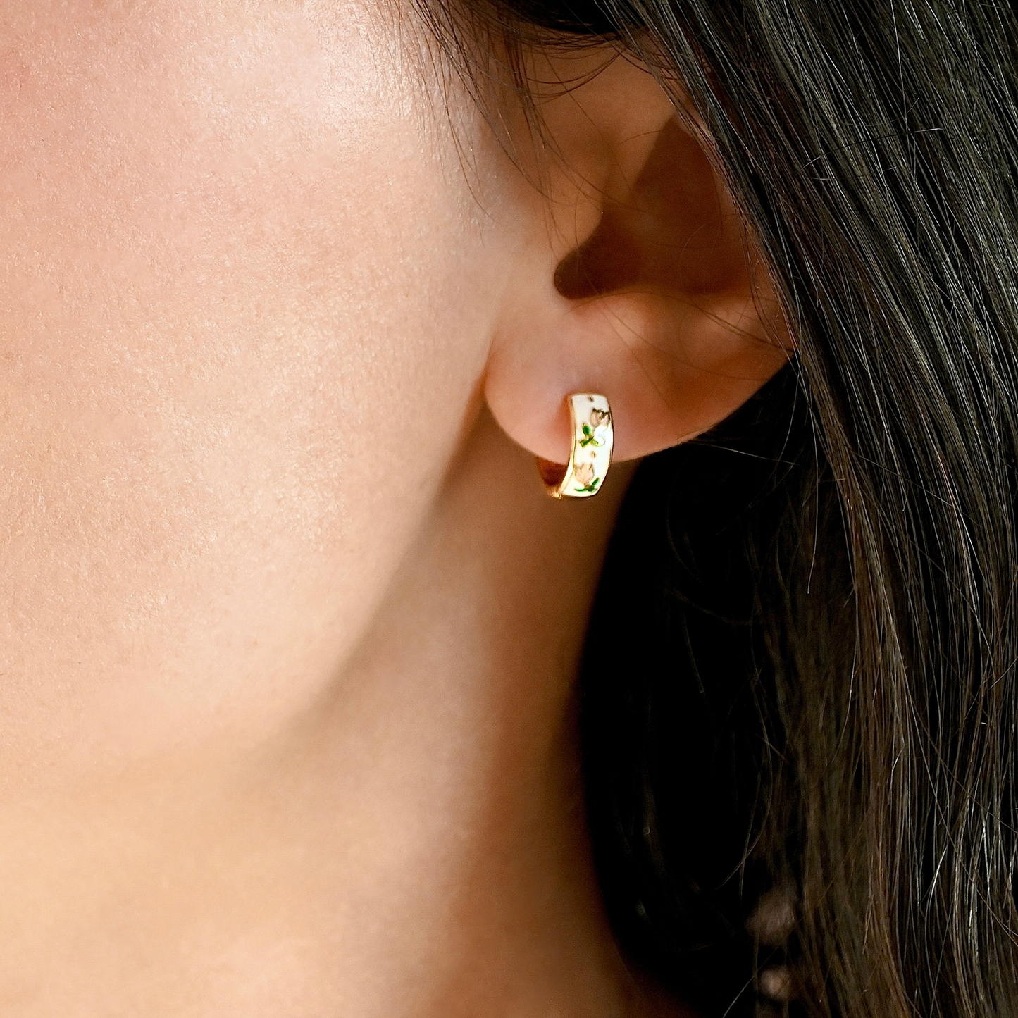 A close-up of a person's ear wearing a small gold huggie hoop earring with tiny clear stones set around a central gold element.