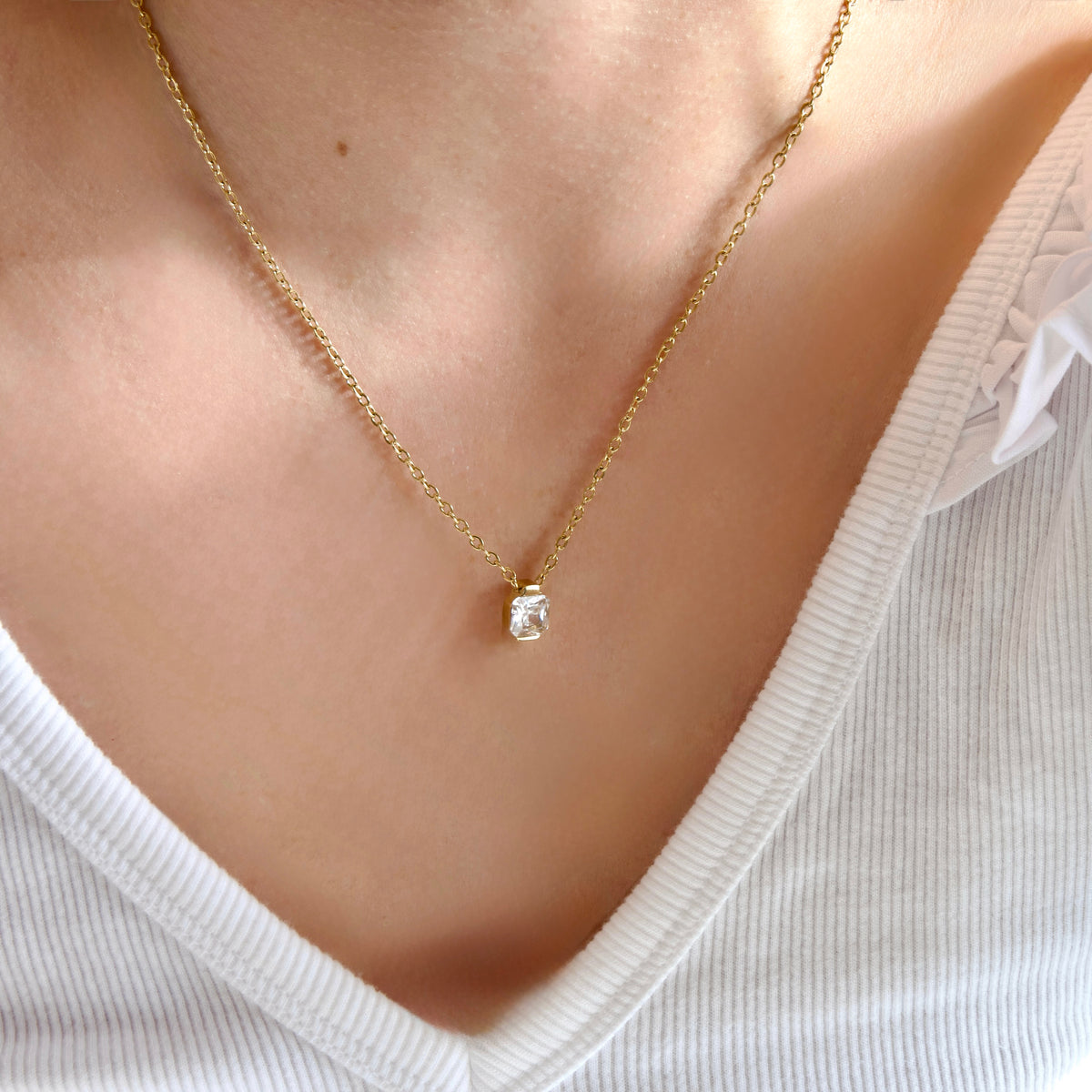 A close-up of a person's neck and chest wearing a delicate gold chain necklace with a small bezel-set clear stone pendant, paired with a white ribbed V-neck top.