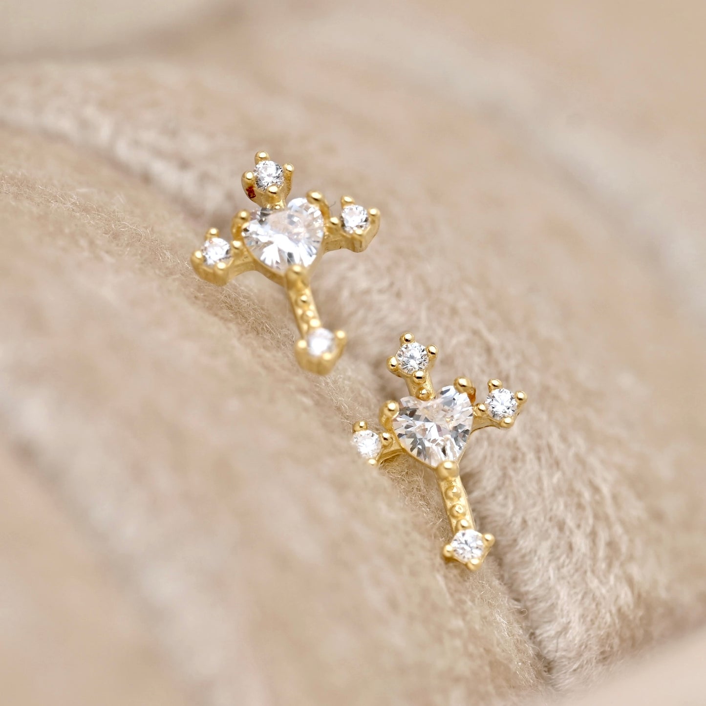 A pair of gold stud earrings with heart-shaped clear gemstones and small clear accent stones, displayed on a soft beige textured surface.