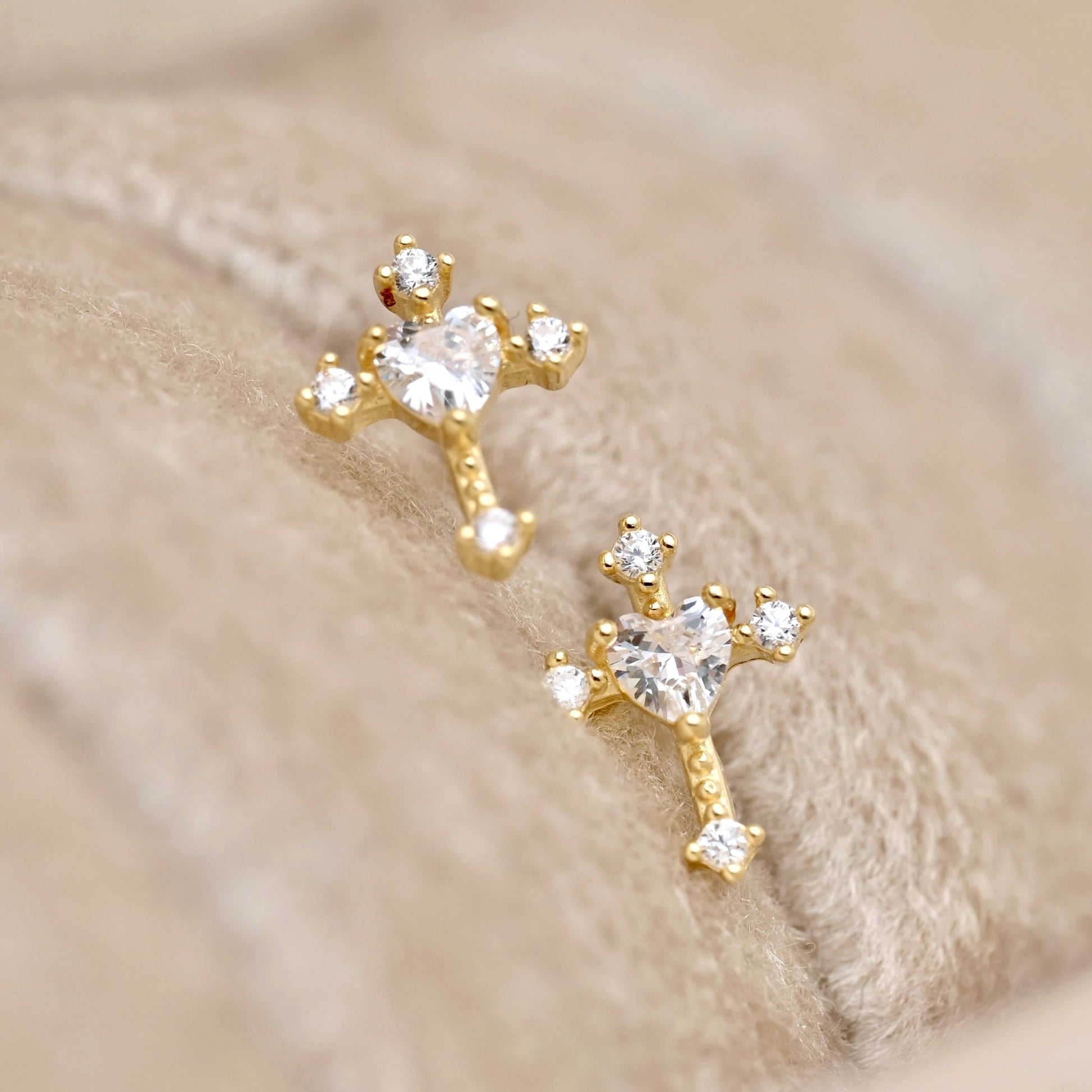 A pair of gold stud earrings with heart-shaped clear gemstones and small clear accent stones, displayed on a soft beige textured surface.