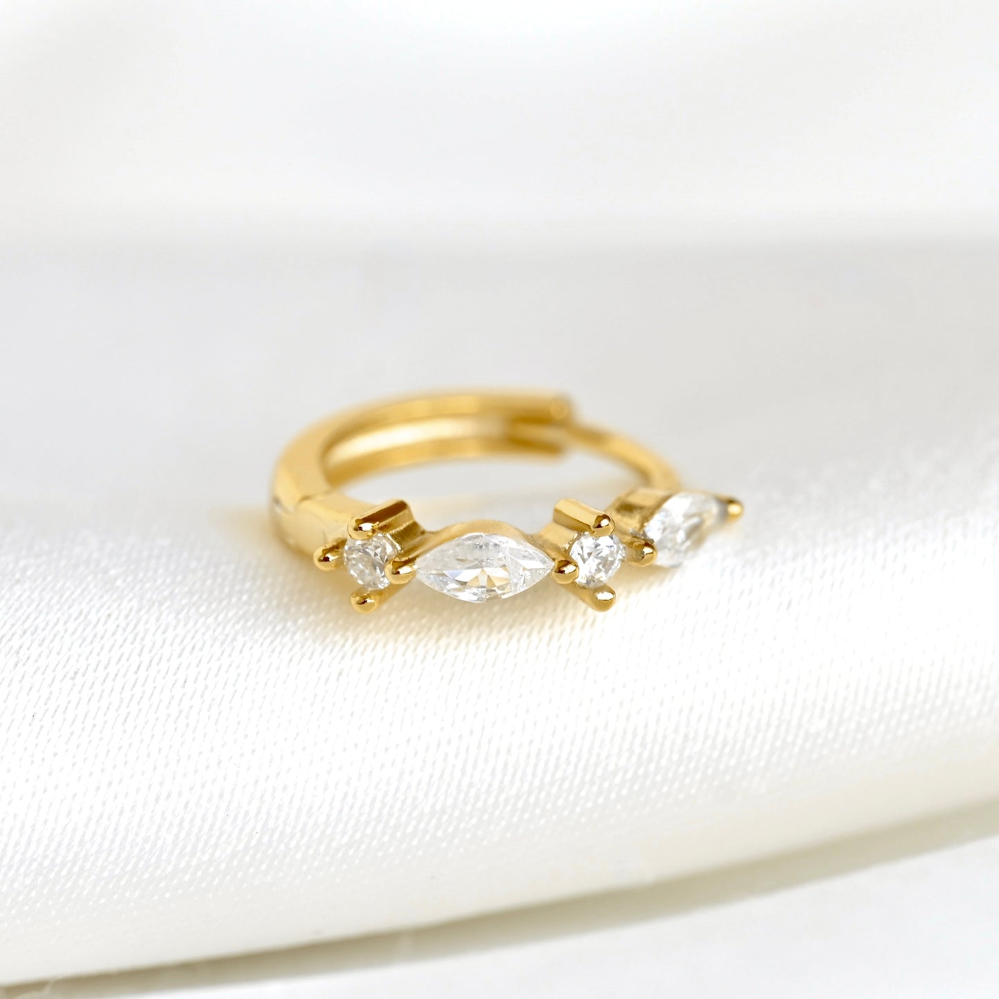 A gold huggie hoop ring featuring a marquise-cut center stone and two small round stones on each side, displayed on a soft white fabric surface.