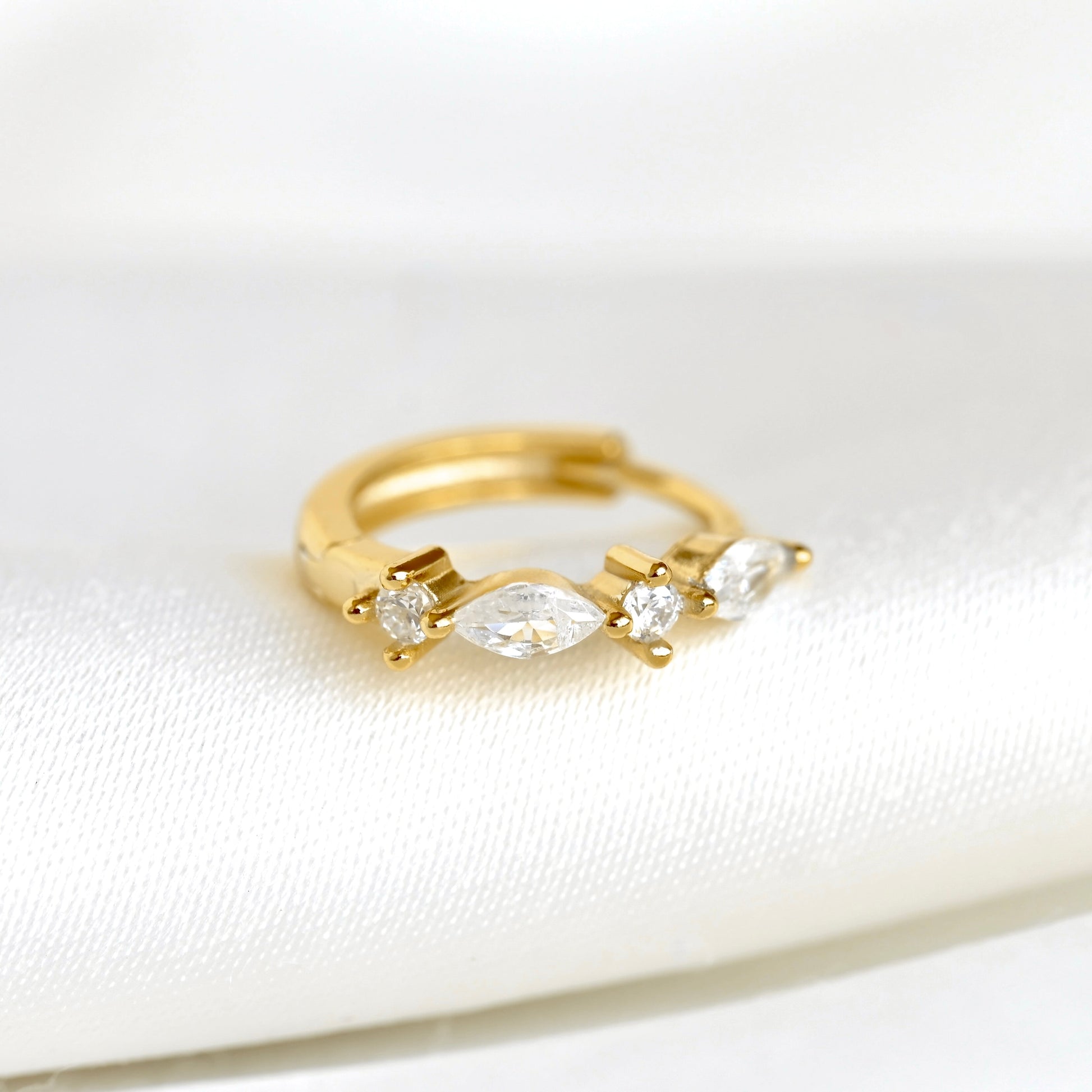 A gold huggie hoop ring featuring a marquise-cut center stone and two small round stones on each side, displayed on a soft white fabric surface.