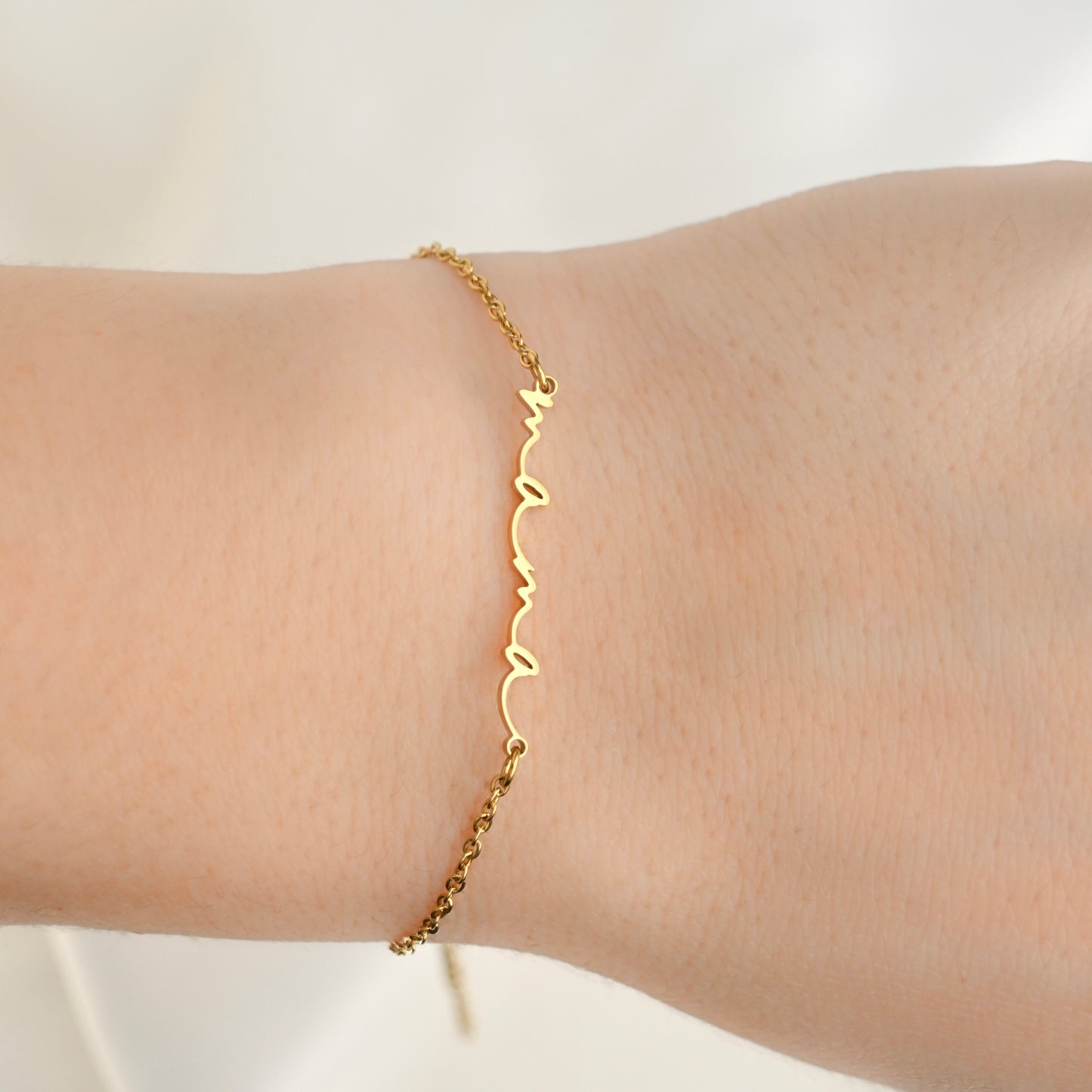 A delicate gold cable chain bracelet with a vertical script nameplate spelling 'mama' in elegant cursive font, shown worn on a person's wrist.