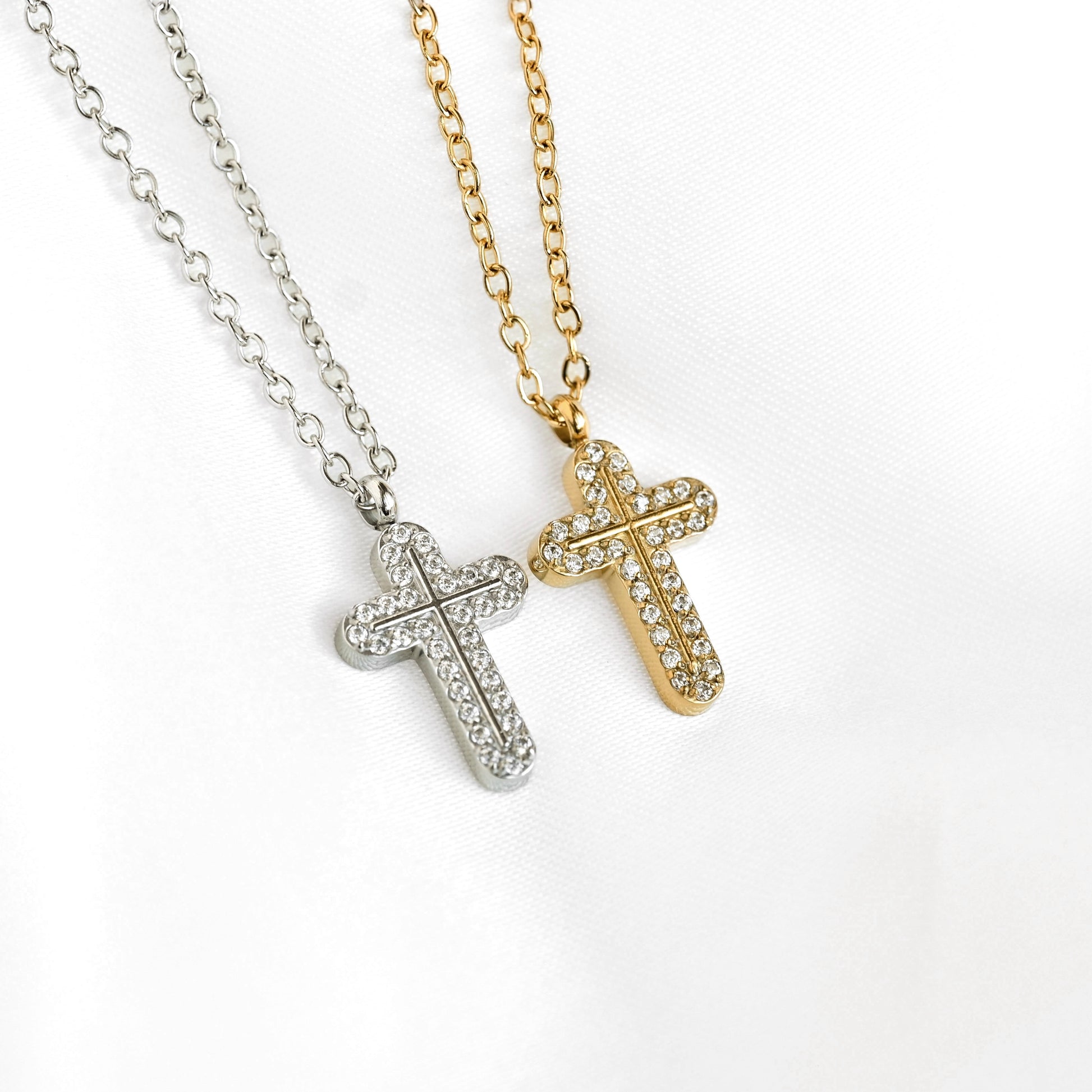 Two cross pendant necklaces on delicate chains, one silver-toned and one gold-toned, both adorned with small clear stones, displayed on a clean white background.