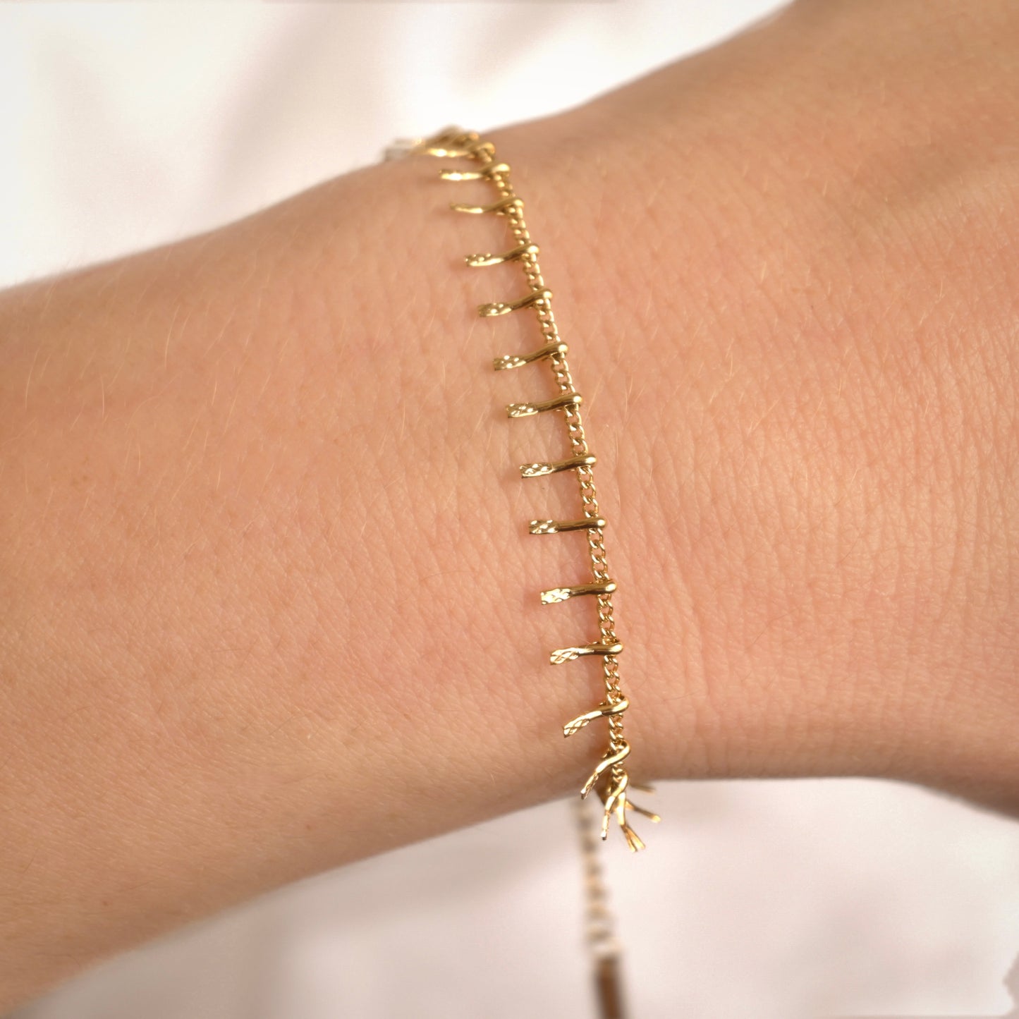 A delicate gold chain bracelet with small dangling leaf-like charms, shown worn on a person's wrist against a soft white background.