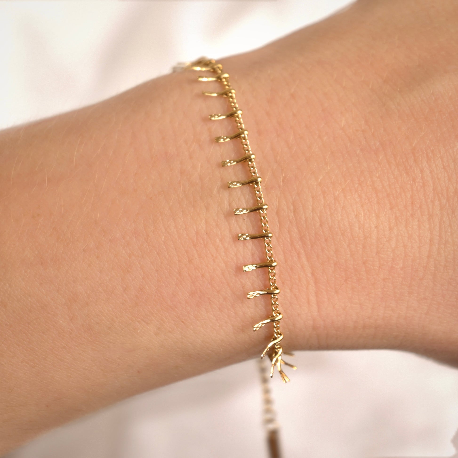 A delicate gold chain bracelet with small dangling leaf-like charms, shown worn on a person's wrist against a soft white background.