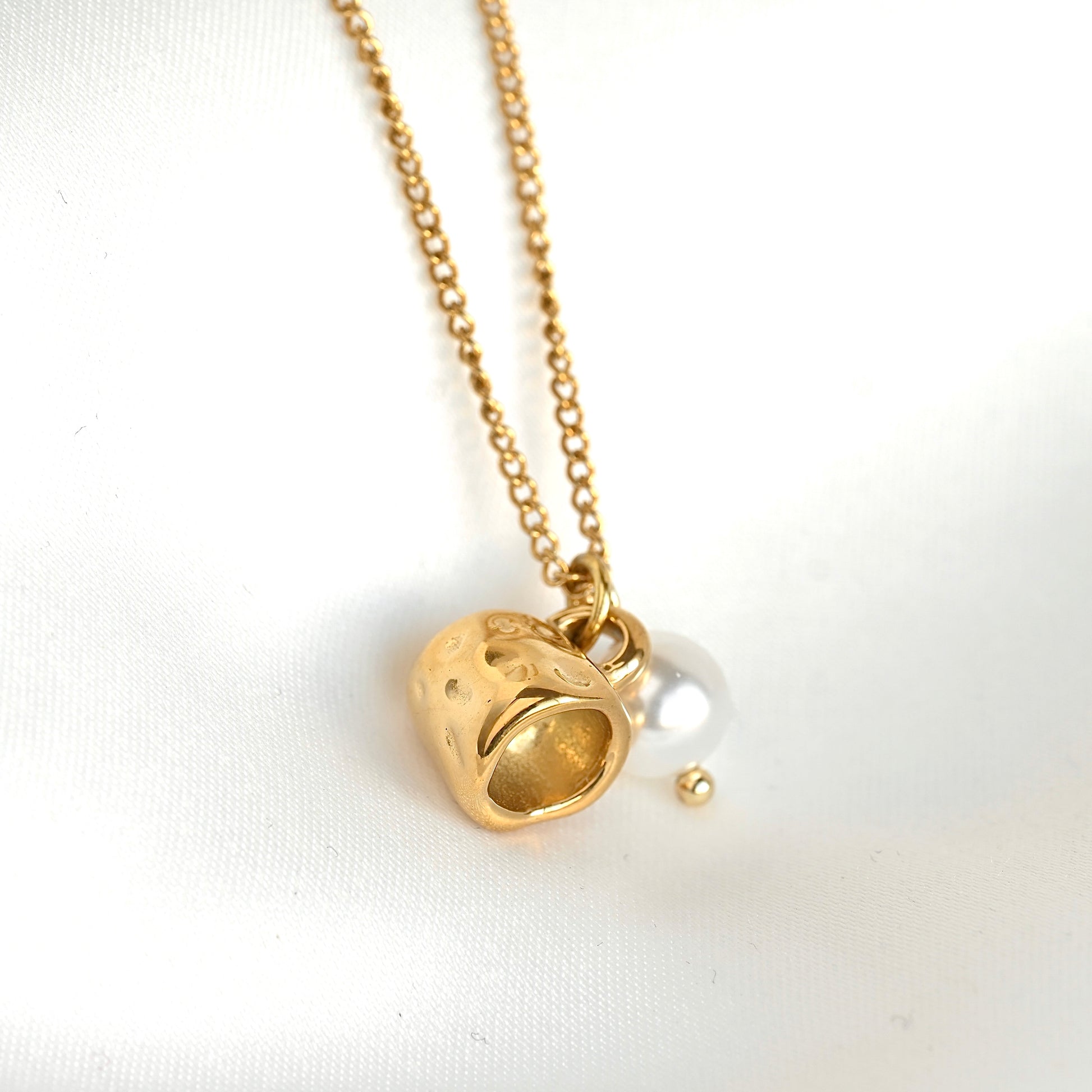 A gold chain necklace featuring a textured gold mug pendant and a small white pearl bead, displayed on a white fabric background.