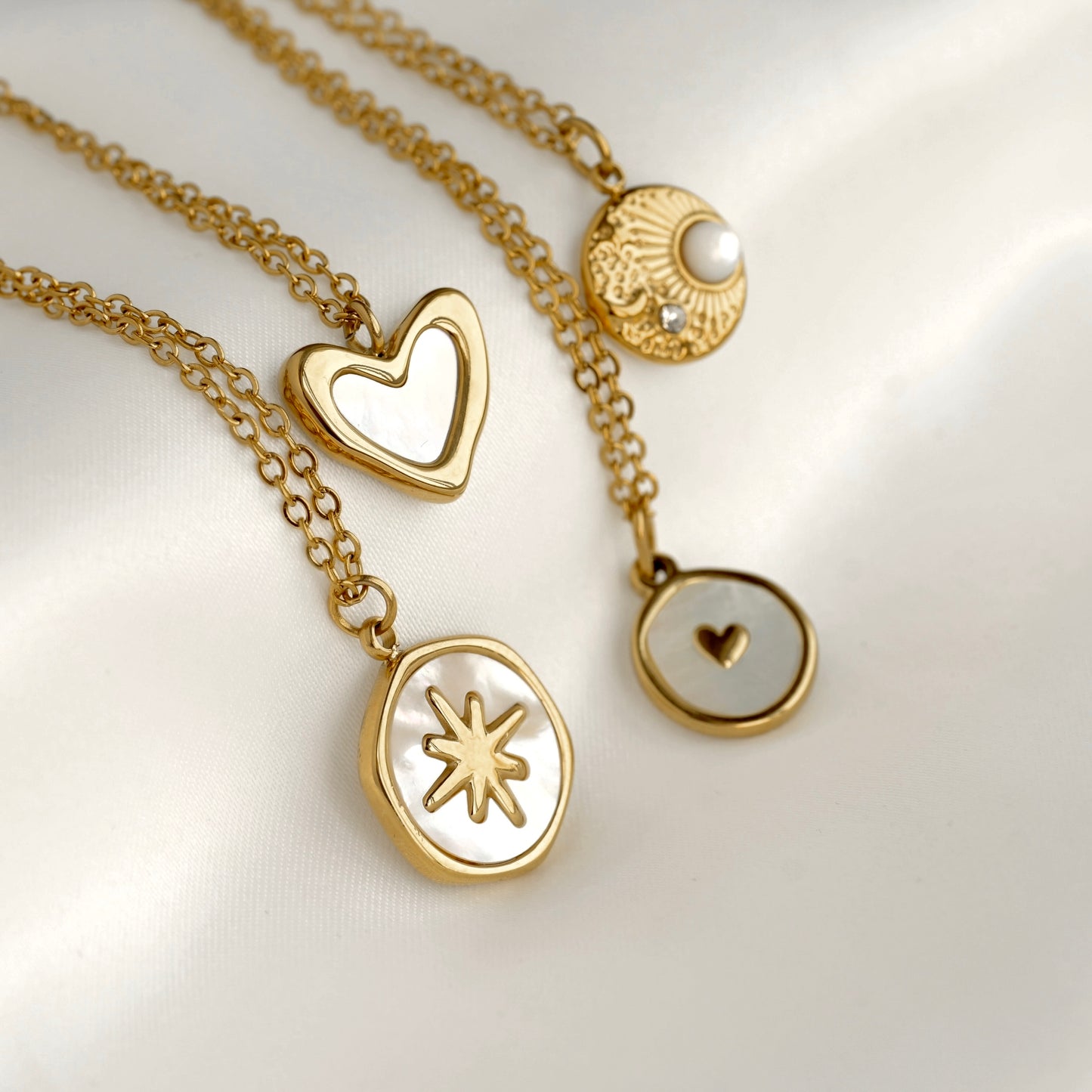 Four gold-tone pendant necklaces arranged on a soft white satin background. The necklaces feature a heart-shaped pendant with a white inlay, an oval pendant with a gold starburst motif, and a round pendant with a small white pearl and tiny clear stone.