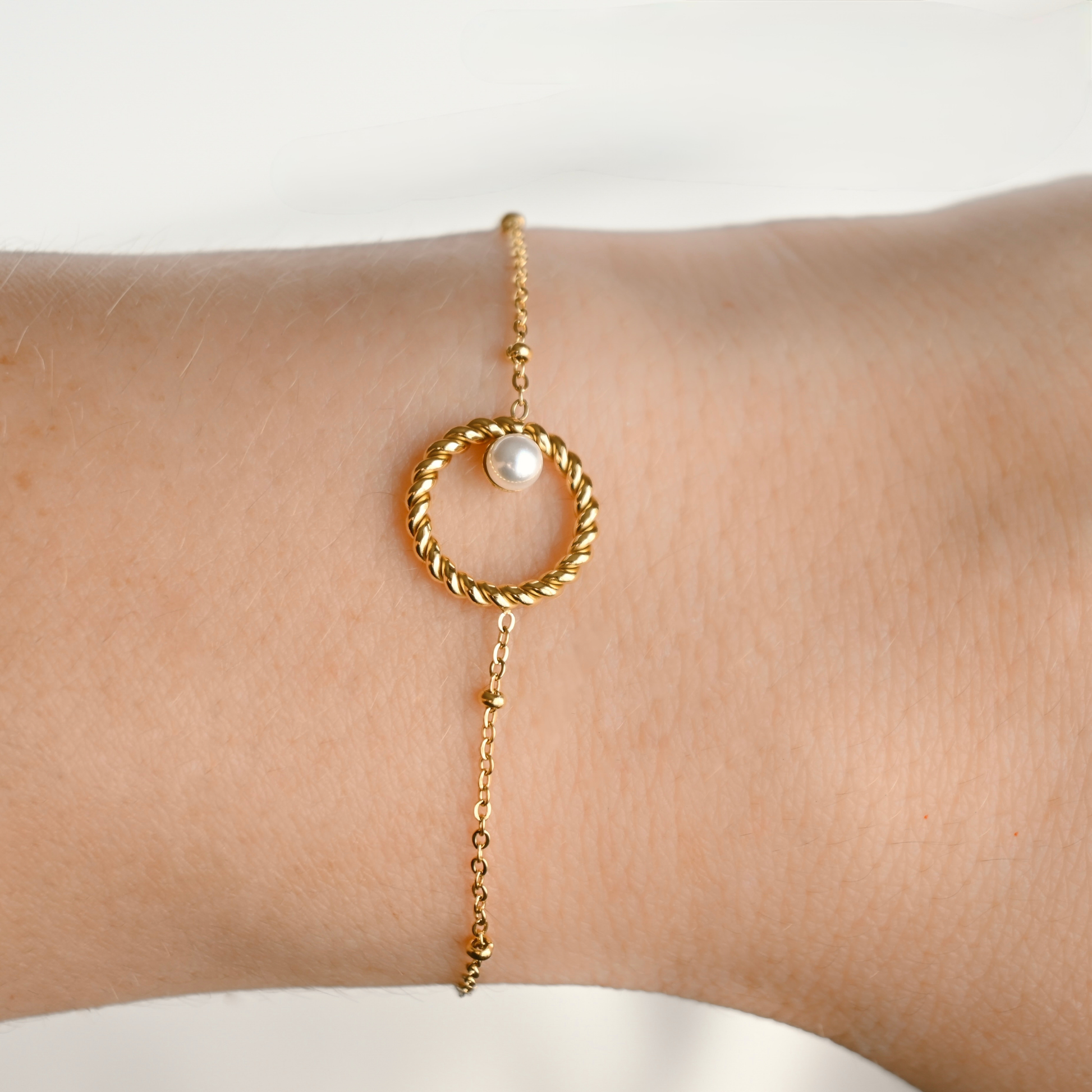 A delicate gold chain bracelet with a twisted circular ring, a small white pearl, and a dangling chain, shown worn on a person's wrist.