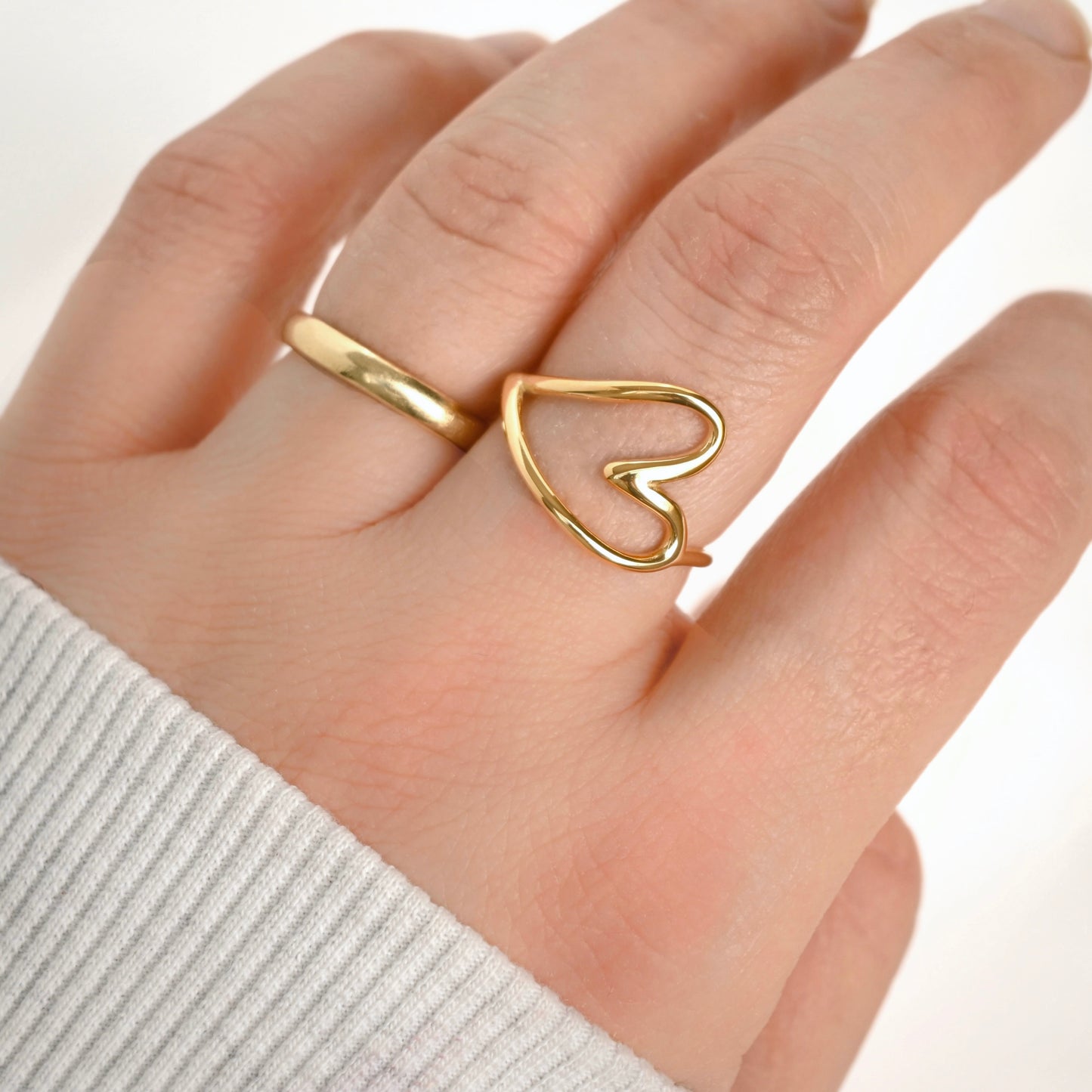 A close-up of a hand wearing two gold rings, with one ring featuring an open heart design and the other a simple polished  wedding band.