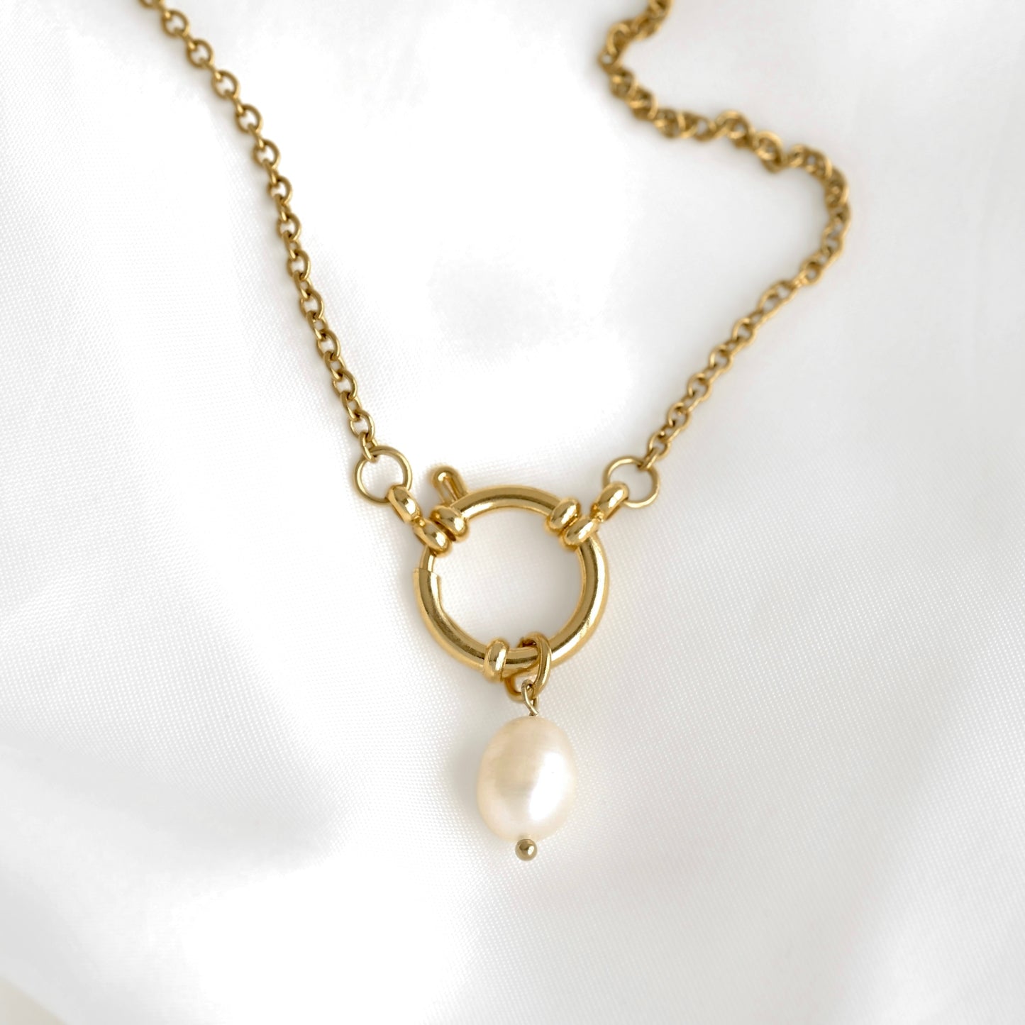 A gold chain necklace featuring a large open circular sailor clasp with a small pearl hanging from its bottom, displayed on a white satin fabric background.