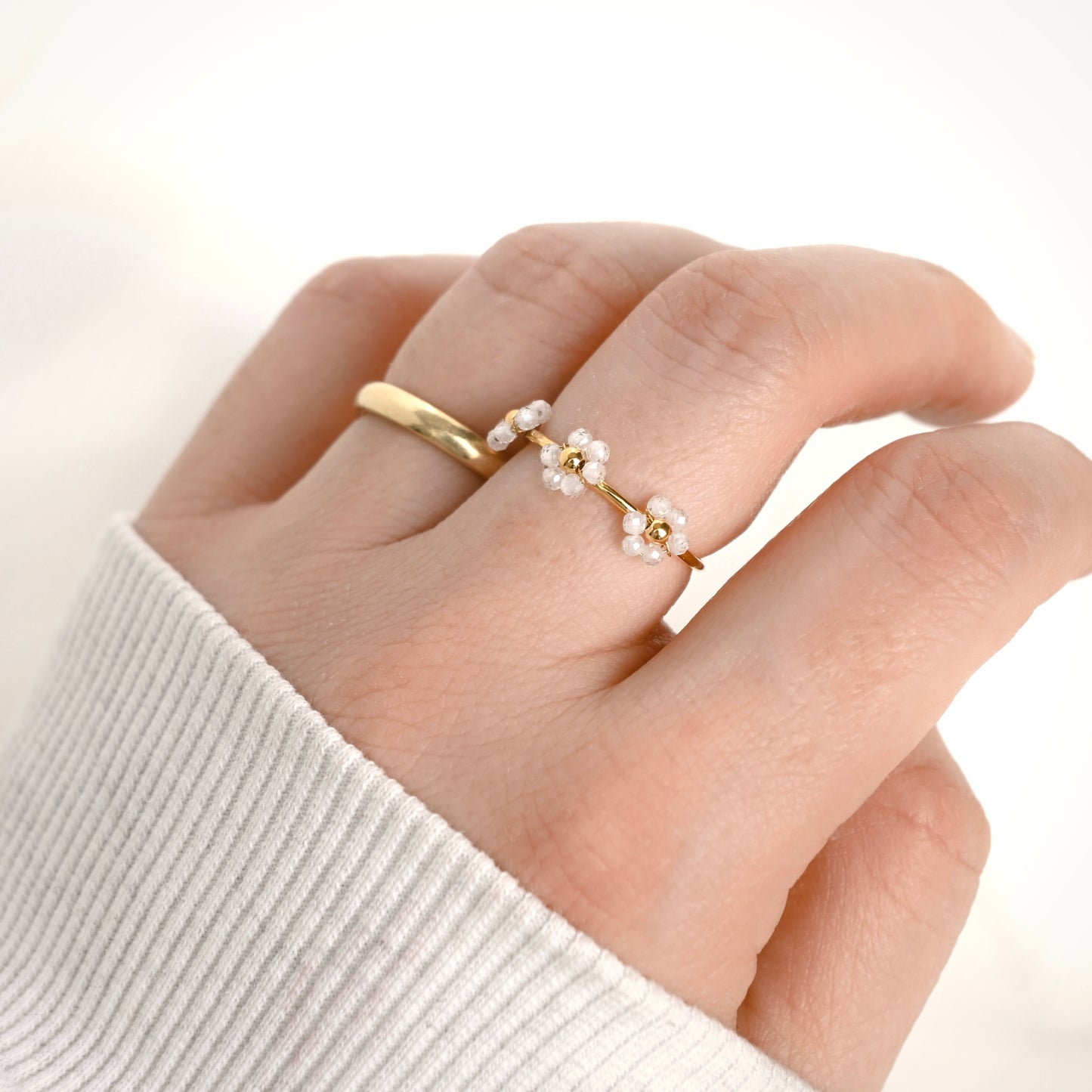 A delicate gold ring featuring three small white beaded flower clusters, shown worn on a finger. The hand is wearing a white ribbed knit sweater cuff, set against a bright, clean, off-white background.