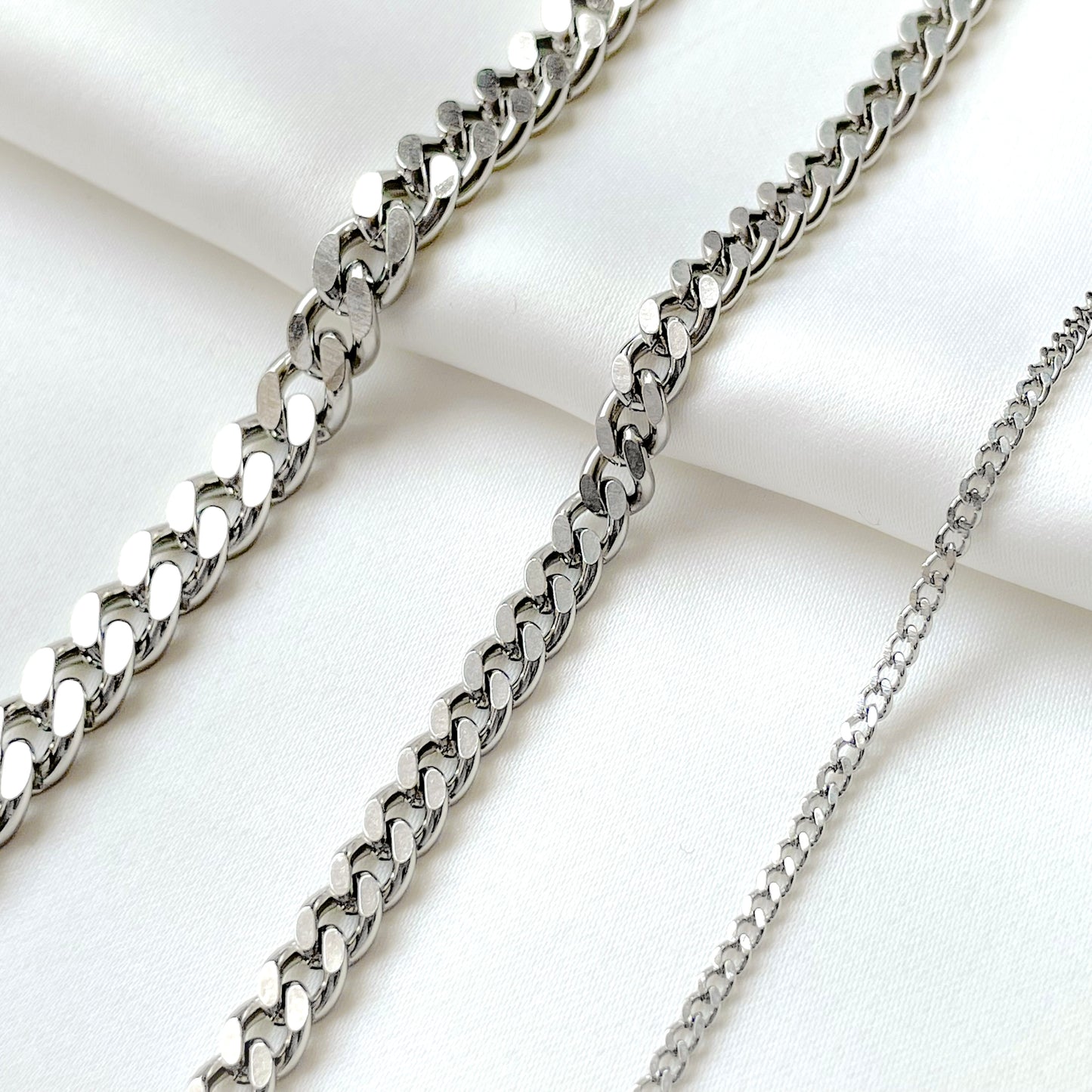 Three silver cuban chains in 2mm, 5mm and 7mm on a white fabric background