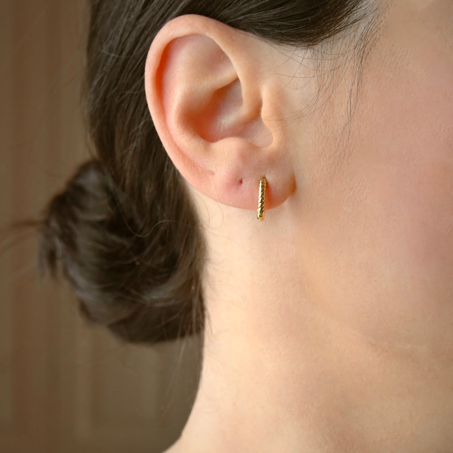 Close-up of an ear wearing a textured gold hoop earring with a blurred background