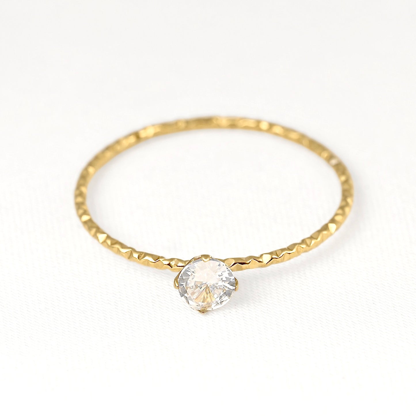 A delicate gold ring with a textured, hammered band and a single clear faceted gemstone in a prong setting, shown on a clean white background.