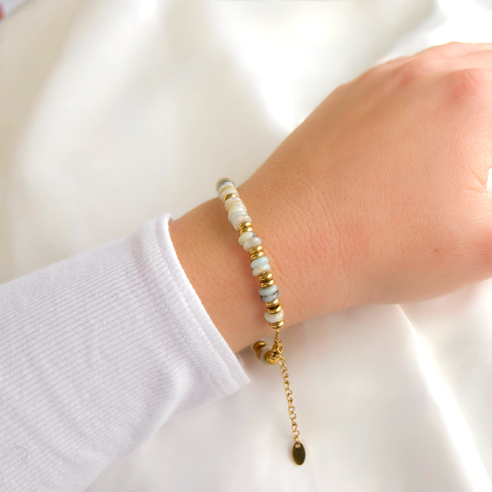 A close-up of a person's wrist wearing a delicate beaded bracelet. The bracelet features light green and white stone beads, small gold spacer beads, and a gold clasp with an adjustable chain and teardrop charm.