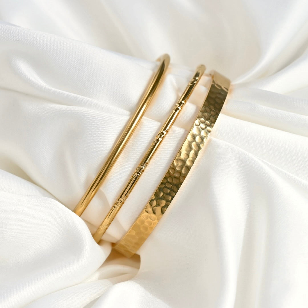 Three gold-toned cuff bracelets displayed on draped white satin fabric. One bracelet is smooth and polished, while the other two feature textured patterns, including a hammered finish.