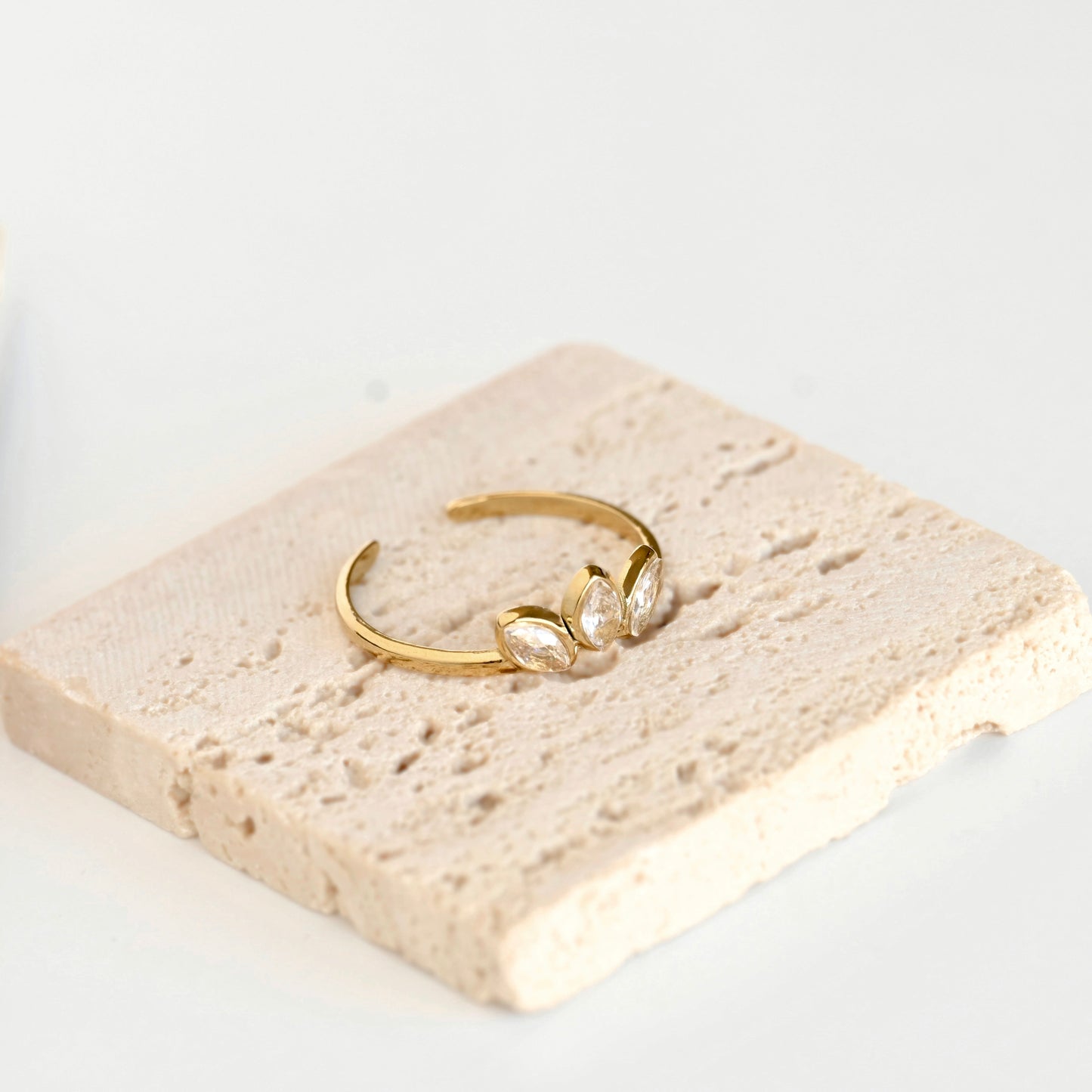 A delicate gold open ring with three marquise-cut clear stones, displayed on a textured beige stone tile against a clean white background.