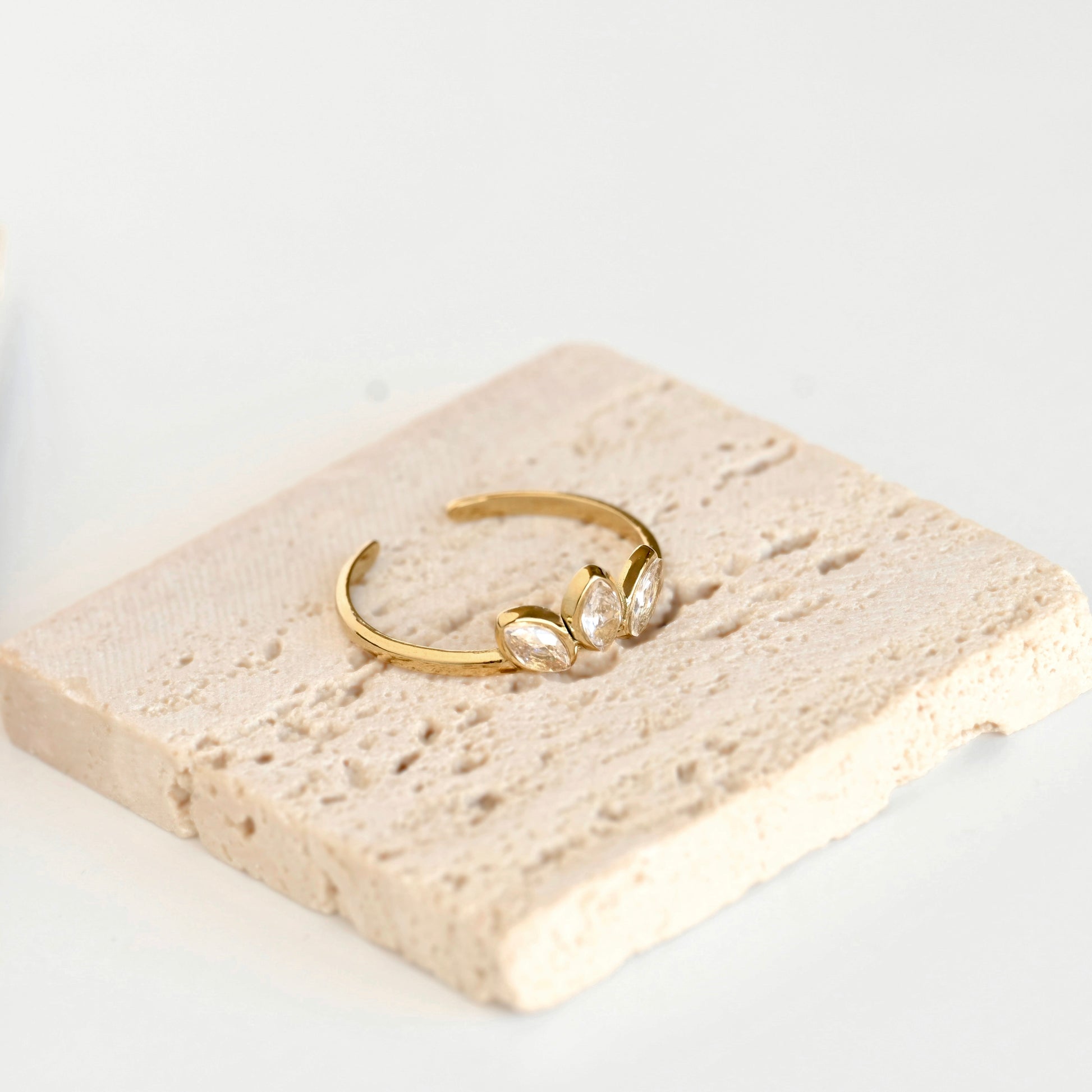 A delicate gold open ring with three marquise-cut clear stones, displayed on a textured beige stone tile against a clean white background.