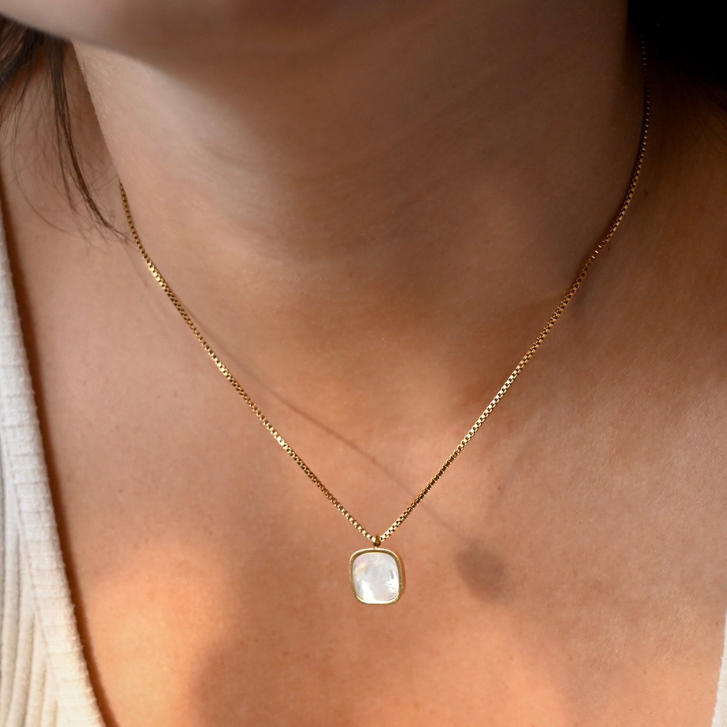 A close-up of a person's neck wearing a delicate gold chain necklace with a small square pendant featuring a milky white stone.