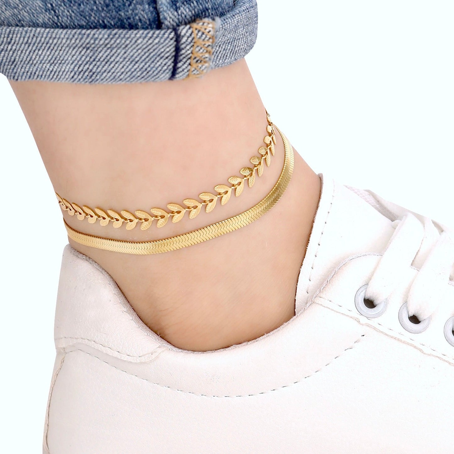 Gold anklets on a person's ankle with a white sneaker and denim jeans in the background.