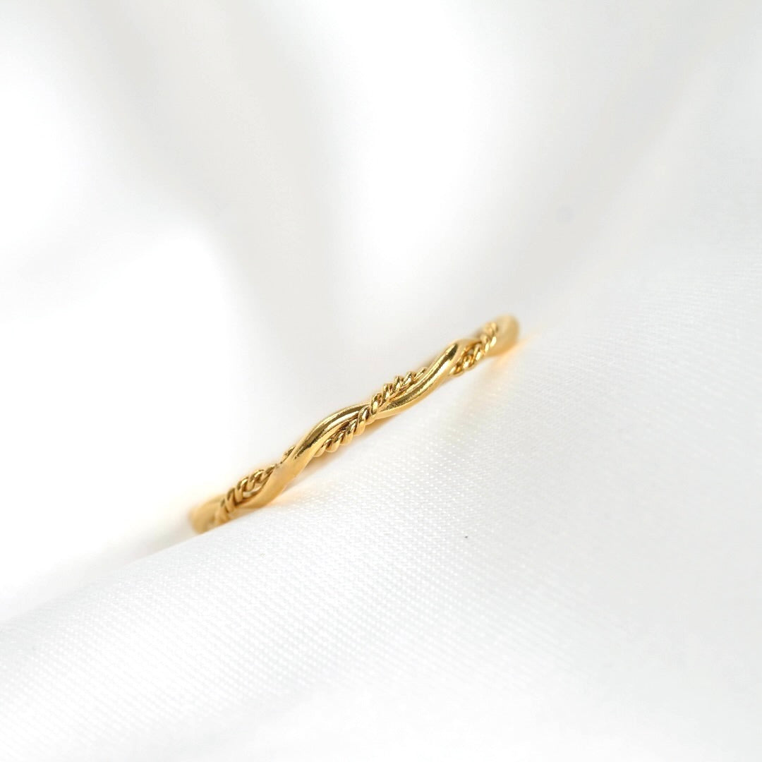 A delicate gold ring with a twisted rope or vine pattern, resting on a soft, white textured fabric background.