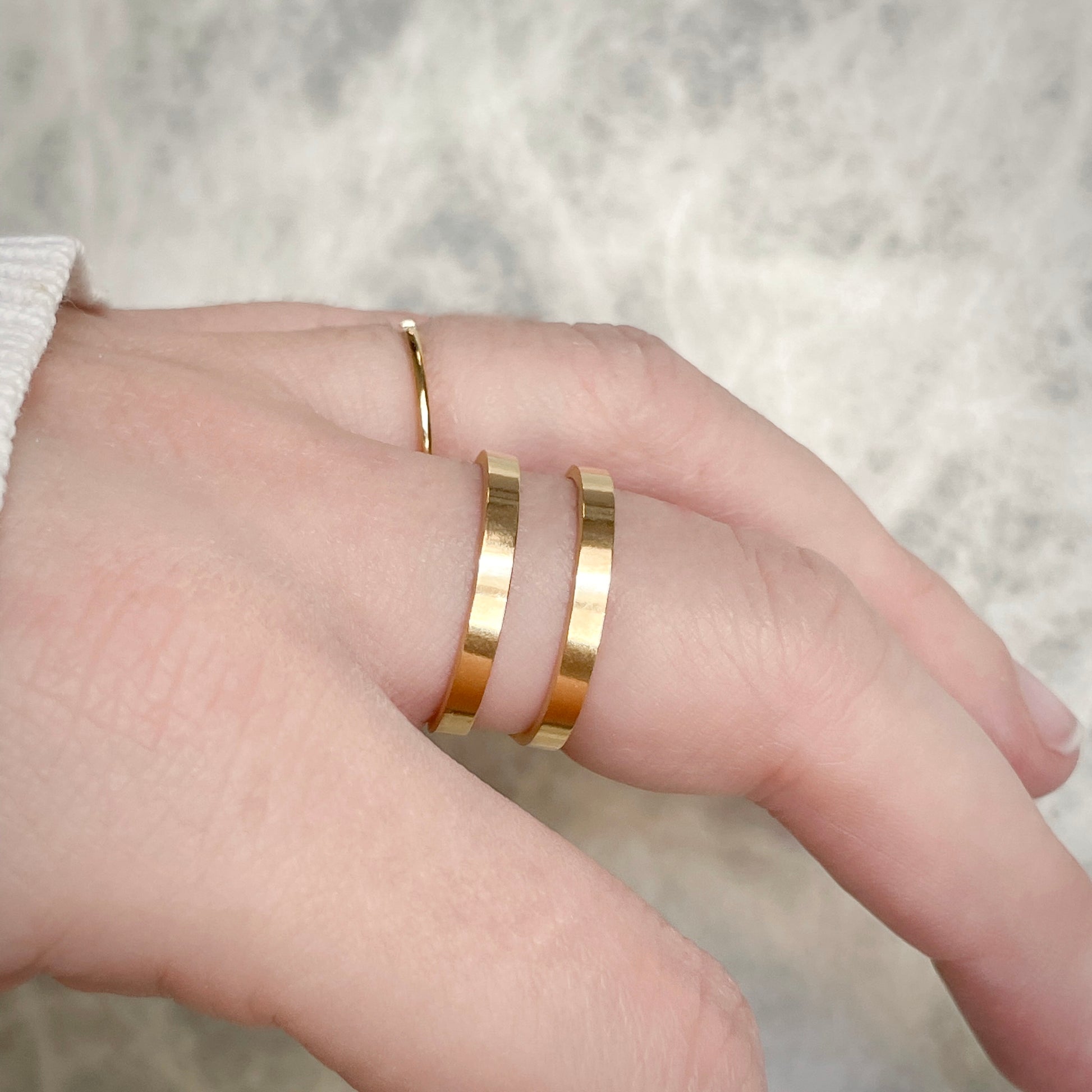 Side view of hand wearing a double layered gold ring with the flat surface of bands reflecting light against a neutral background