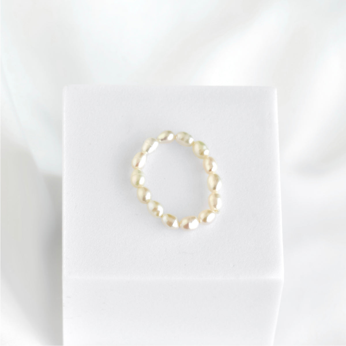 A delicate beaded ring with small, irregular cream-colored beads, displayed on a white jewelry stand against a soft white fabric background.
