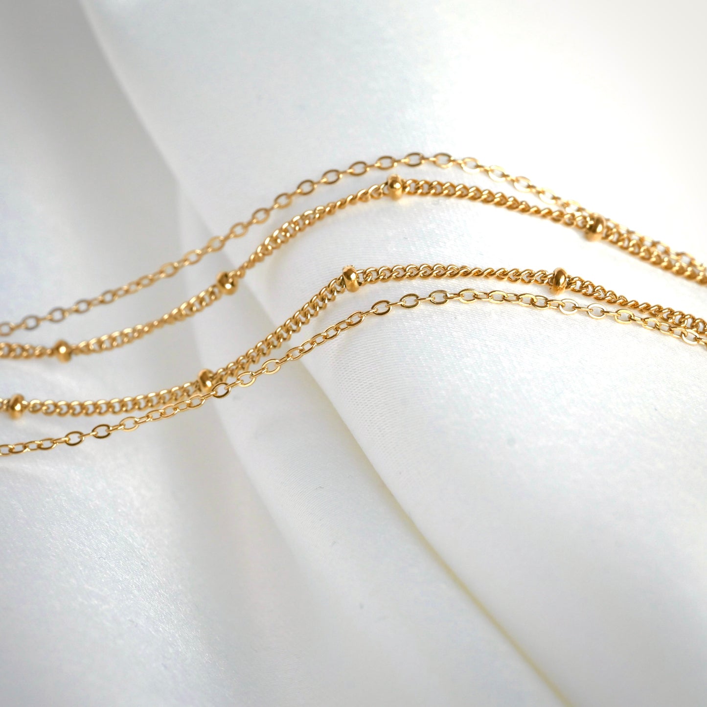 Two delicate gold chains,  one cable and one beaded satellite, on a white fabric background