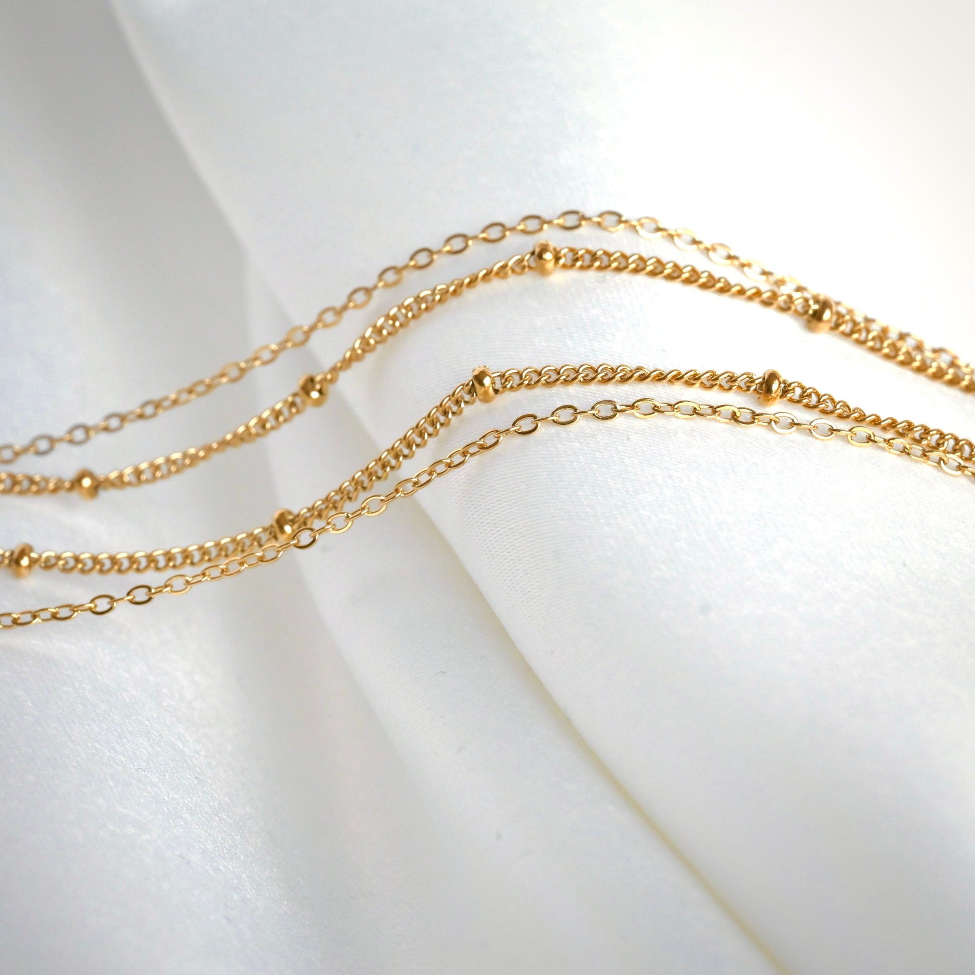 Two delicate gold chains,  one cable and one beaded satellite, on a white fabric background