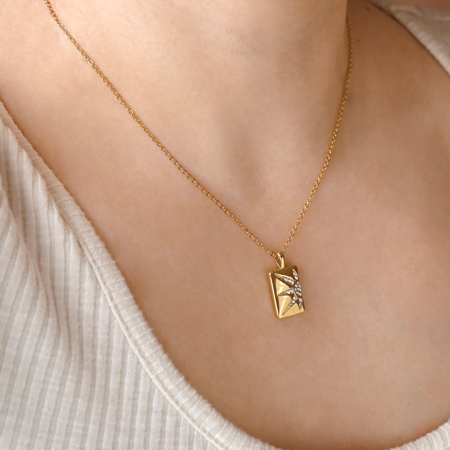 A close-up of a person's neck and chest wearing a delicate gold chain necklace with a small rectangular pendant featuring a starburst design with clear stones.