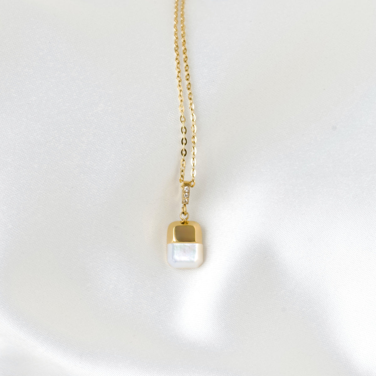 Front view of a delicate gold chain necklace with a small gold cap and a white pearlescent rectangular pendant, displayed on a soft white satin surface.