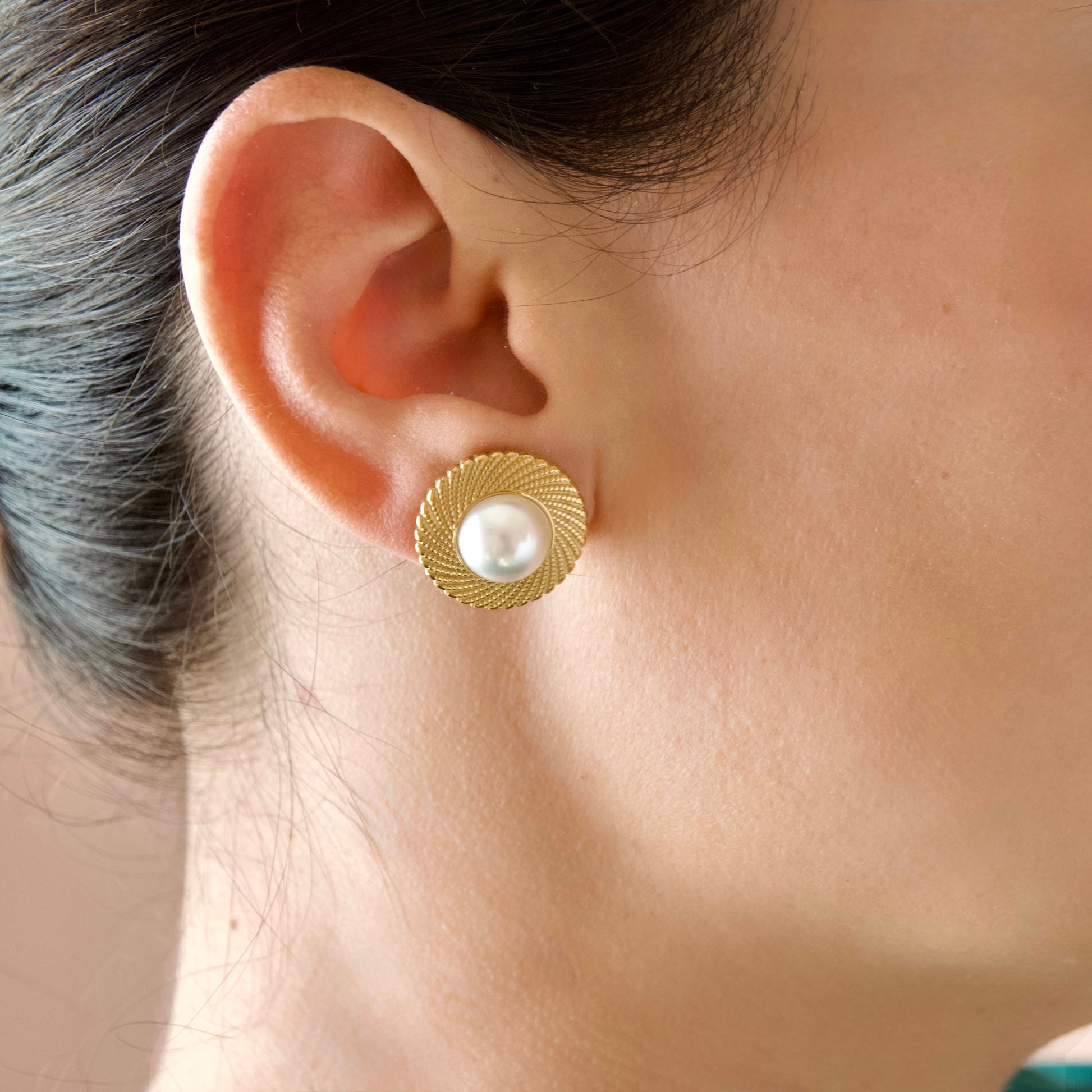 A close-up side profile of a woman wearing a gold textured circular stud earring with a white pearl center.